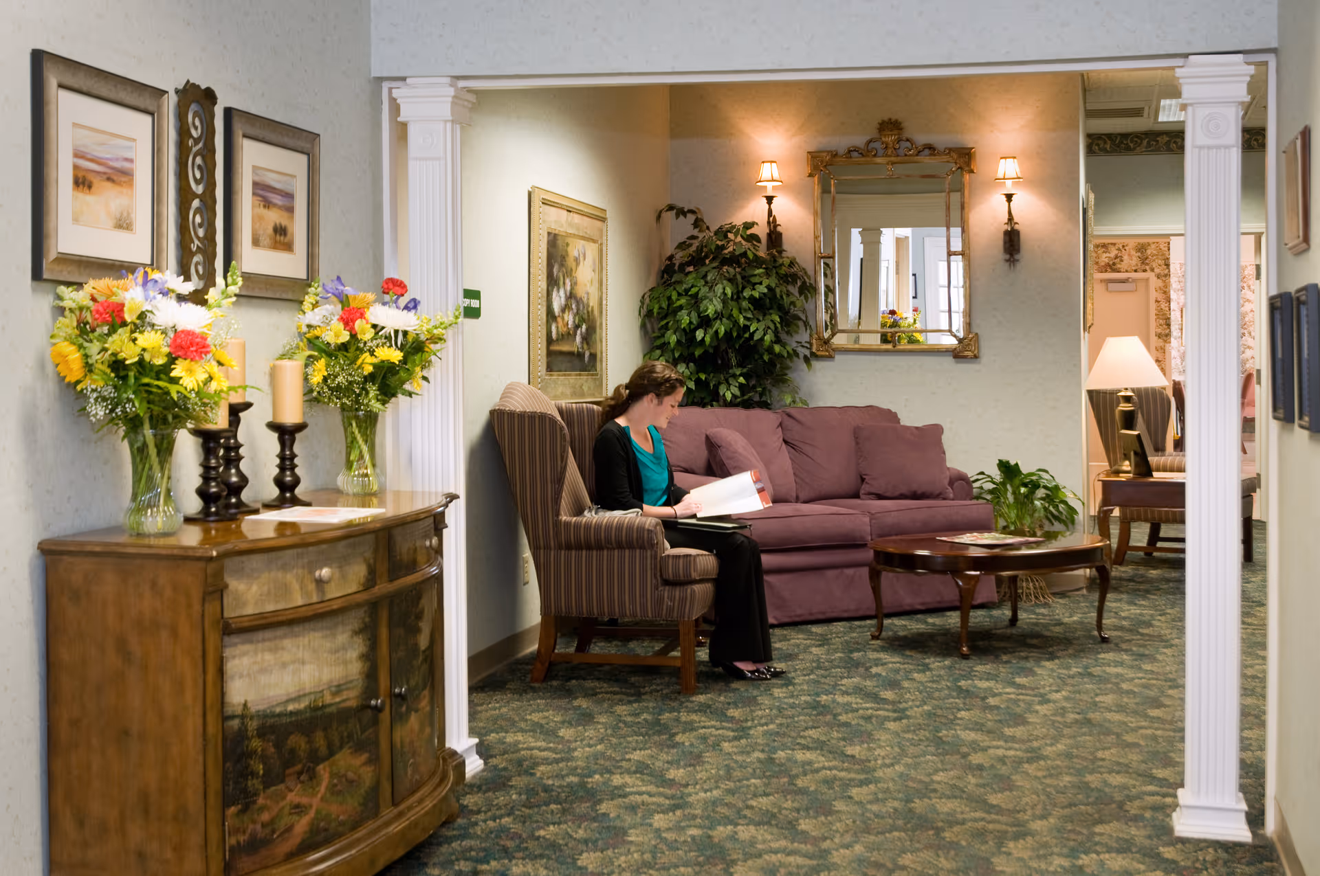 A cozy interior sitting area showing a person reading in an armchair beside a purple sofa, coffee table, plants, framed art, and floral arrangements on a wooden console.