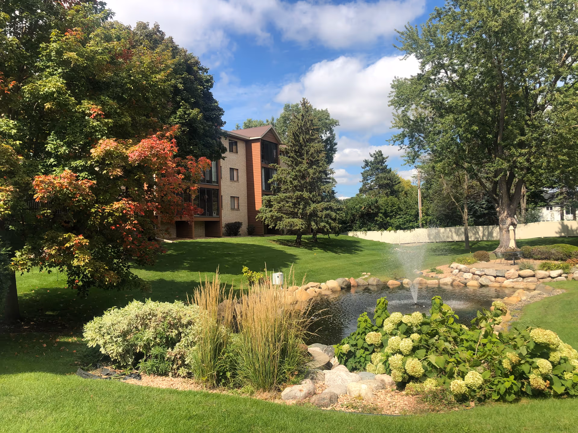 Exterior view of a senior living campus with a pond, fountain, trees, and a landscaped lawn.