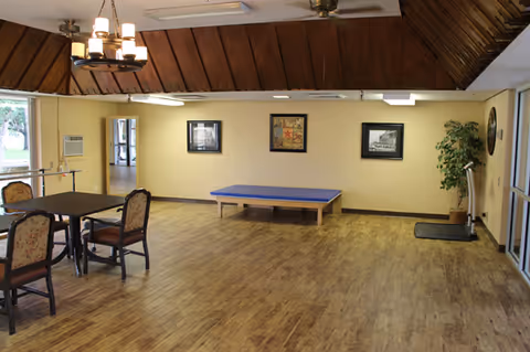 A spacious common recreation room with tables, chairs, a blue activity table, and wall art.