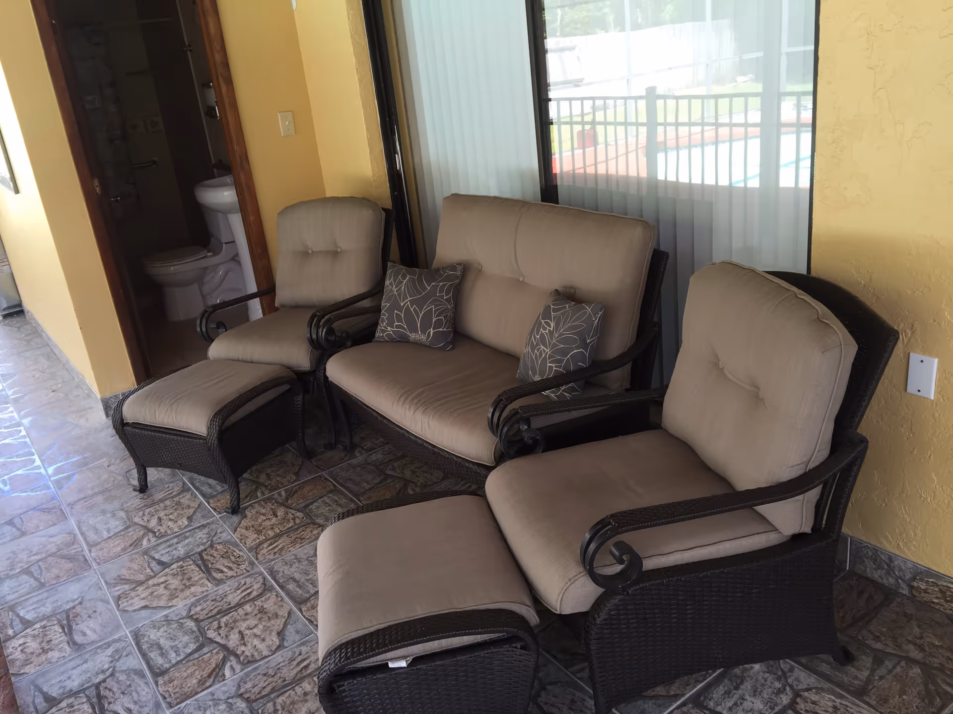 Outdoor patio area with cushioned wicker furniture including two armchairs with matching ottomans and a loveseat with two decorative pillows. The patio has tiled flooring and yellow walls. A bathroom with a visible toilet and sink is seen through an open door on the left side.