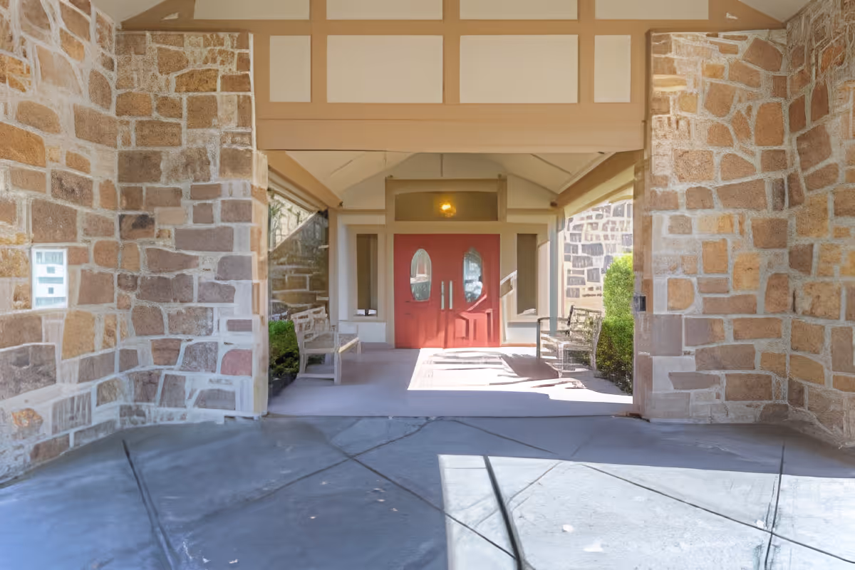 Covered entrance area with stone walls and a concrete floor leading to double red doors with oval windows. There are benches on either side of the walkway and some greenery visible near the walls.