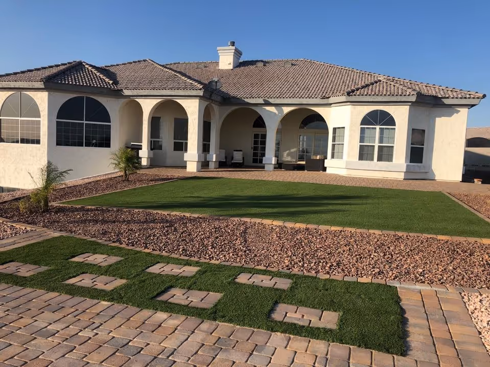 Single-story stucco house with a tiled roof, arched covered patio and a landscaped yard with artificial turf and stone paths under a clear sky.