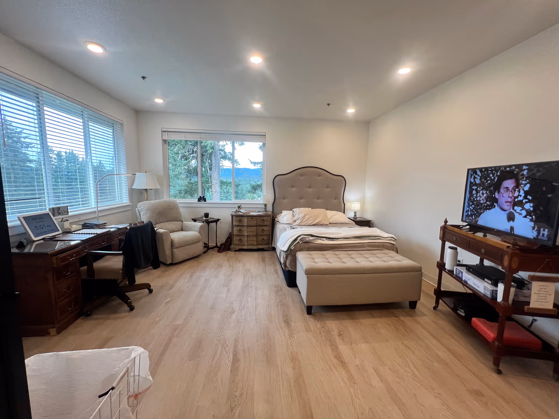 Bright senior living bedroom with bed, ottoman, desk, armchair, TV, and large windows.