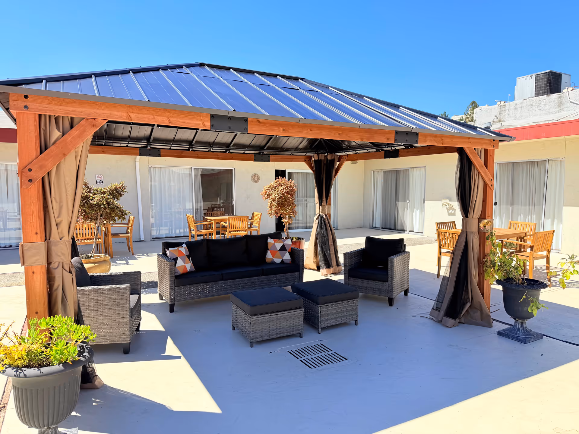 Outdoor patio with wooden pergola, cushioned seating, and dining tables in sunny courtyard.