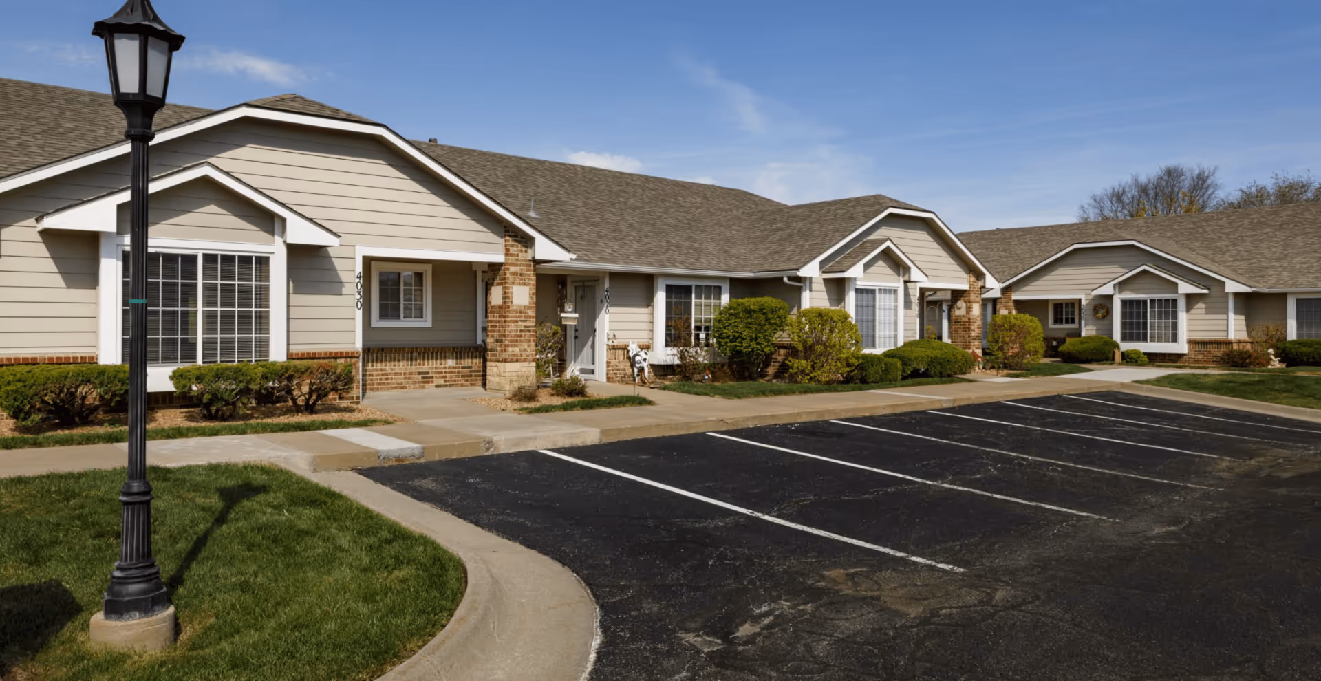 Single-story beige senior living building with brick accents, landscaped shrubs, a lamp post, and an empty parking lot in front.