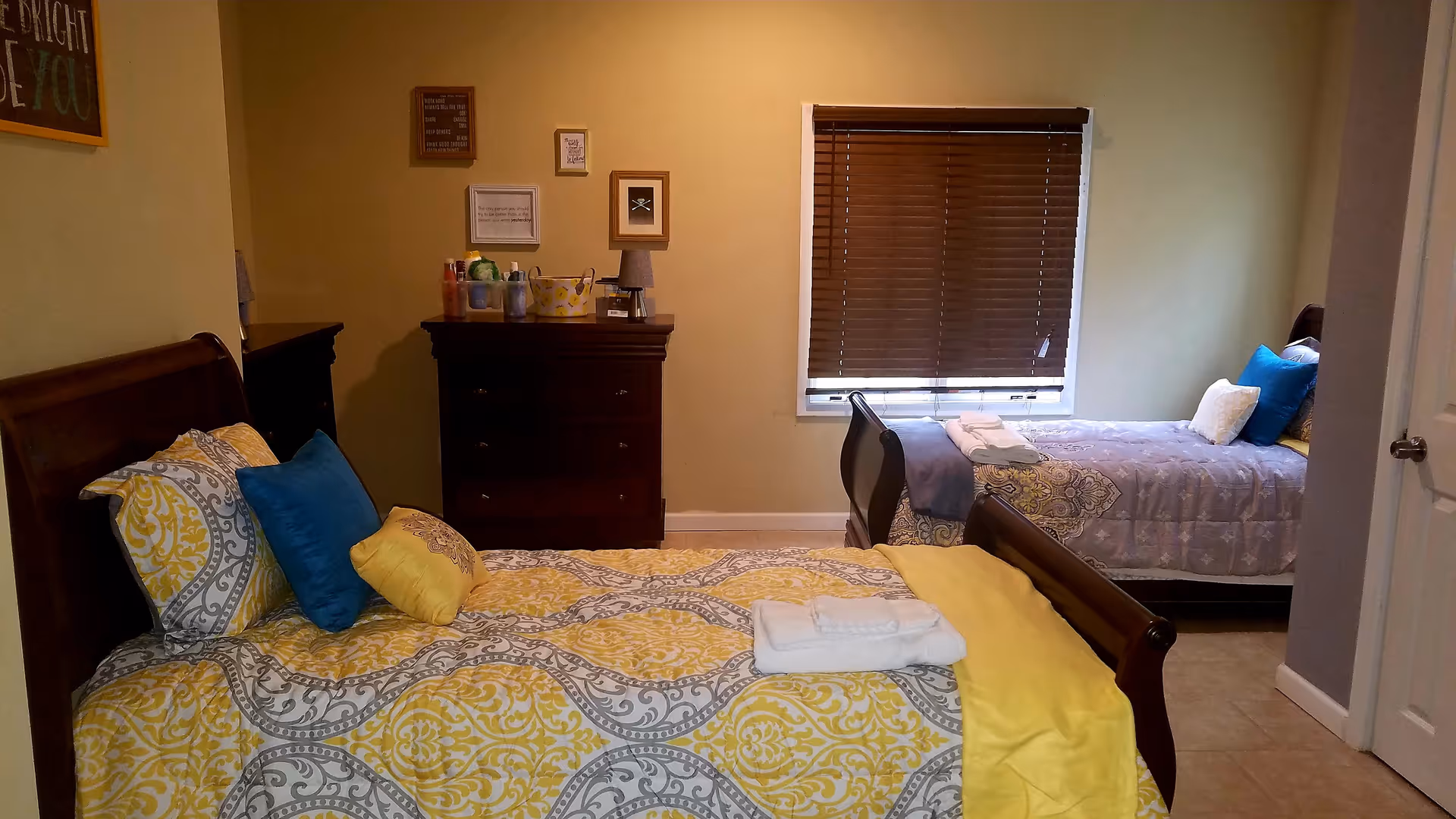 A warmly decorated bedroom with two twin beds, wooden dressers, and folded towels on a bed.