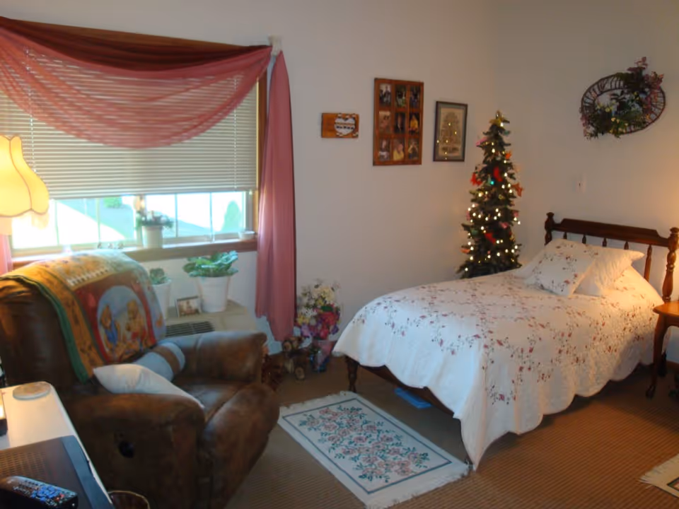 A cozy bedroom with a single bed covered in a white floral quilt, a small decorated Christmas tree with lights next to the bed, a brown recliner chair with a colorful blanket draped over it, a window with pink curtains and blinds, several potted plants on the windowsill, framed pictures on the wall, and a small floral rug on the floor.