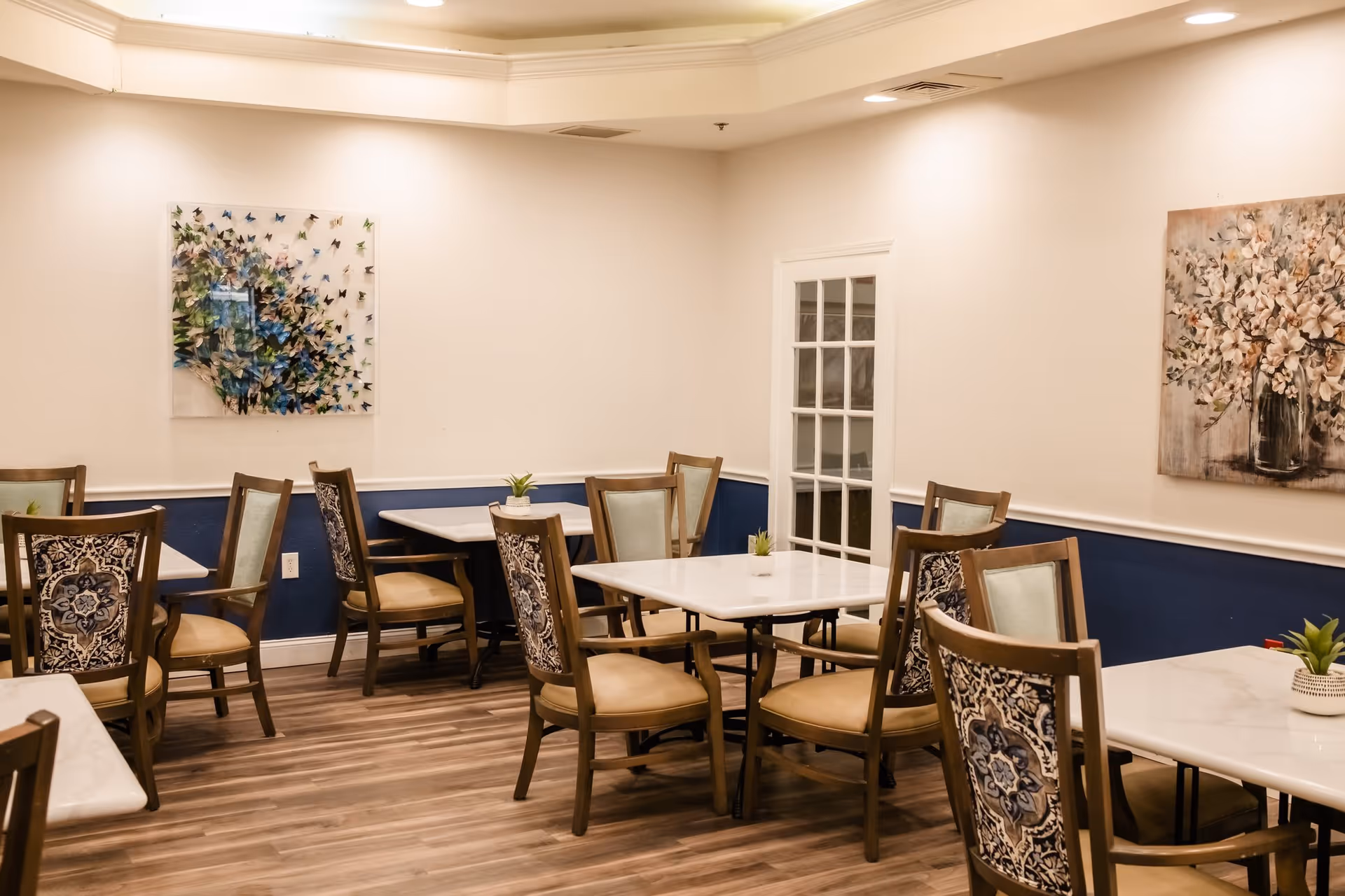 A dining room with several tables and chairs arranged neatly. The chairs have patterned backs and beige cushions. The walls are painted white with a dark blue wainscoting. There are two pieces of artwork on the walls, one featuring a floral design and the other a cluster of butterflies. The floor is wood, and small potted plants are placed on the tables.