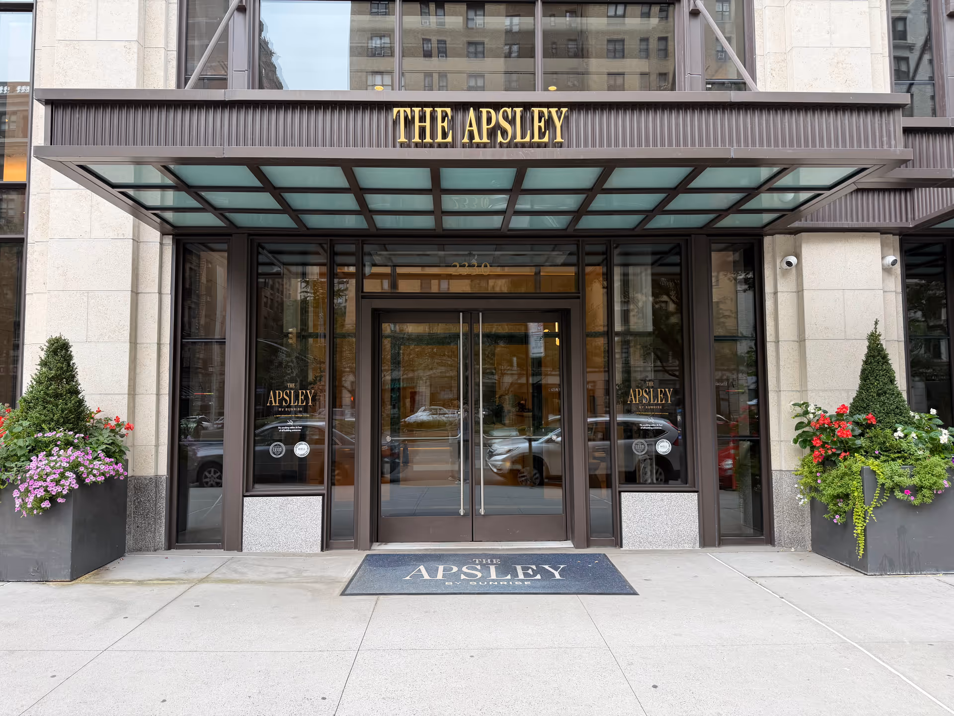 The Apsley building entrance with glass doors, brown frame, and flowering planters on both sides.