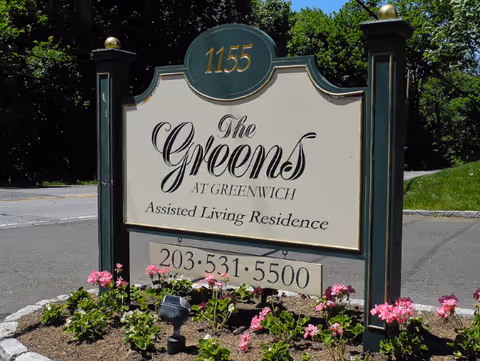 Sign reading The Greens at Greenwich Assisted Living Residence with surrounding pink flowers.