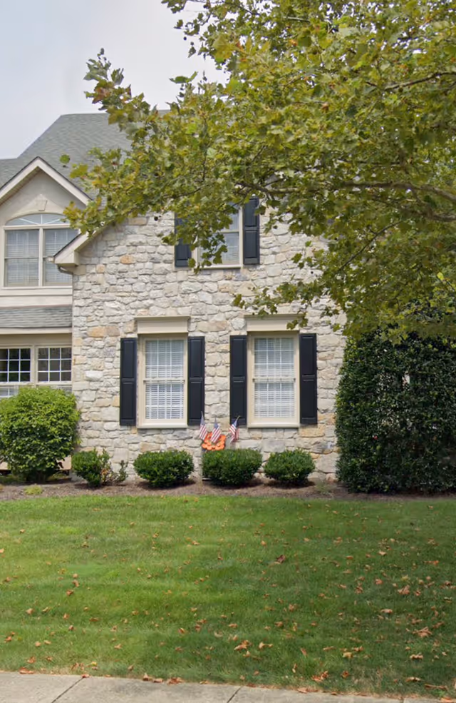 Stone facade senior living building with black shutters, manicured shrubs, and a tree in foreground.