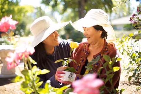 Two women wearing sun hats are gardening together outdoors, smiling and holding a yellow rose plant surrounded by other blooming flowers.