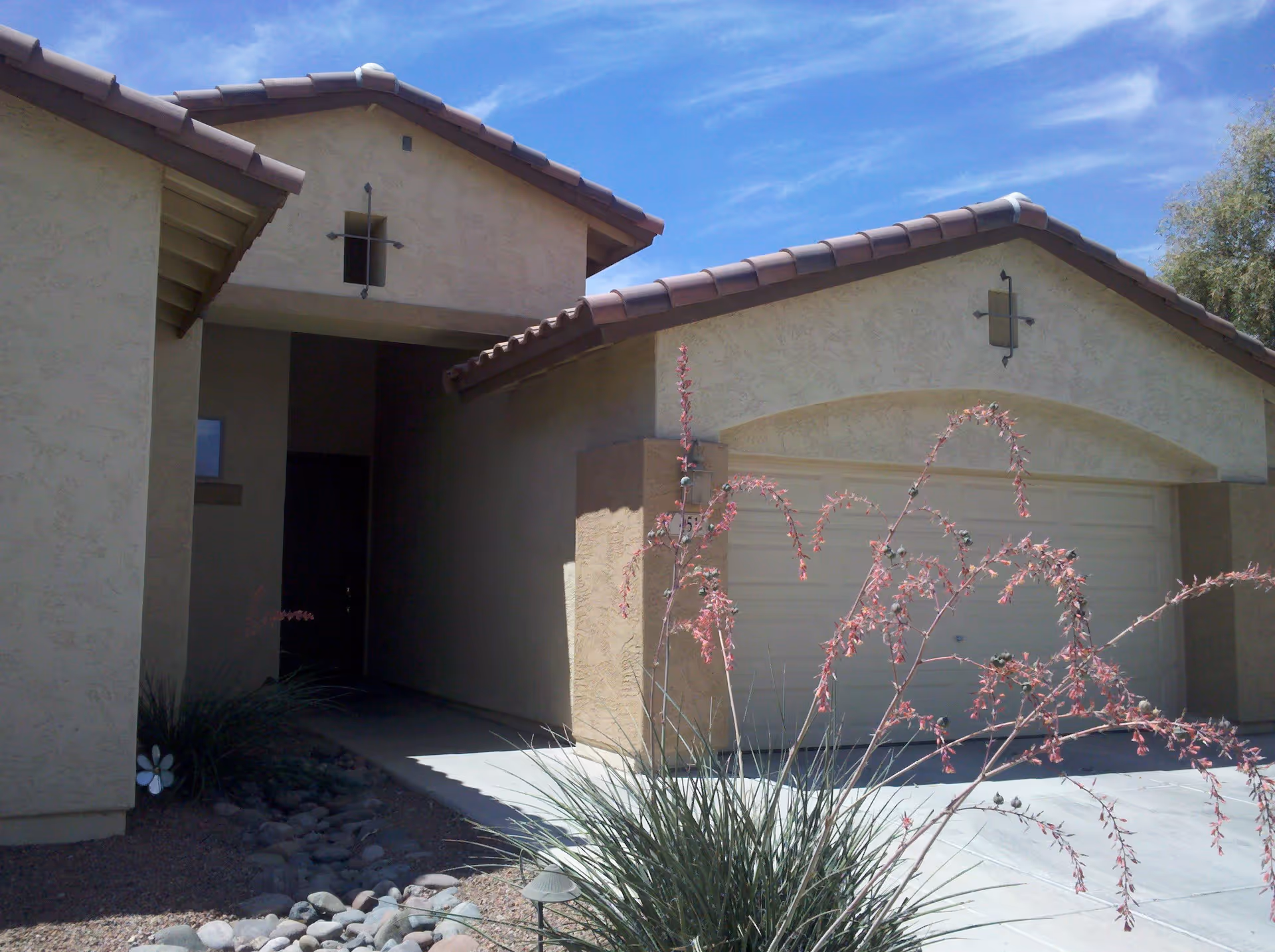 Front exterior of a stucco house with a two-car garage, tiled roof, driveway and desert landscaping.