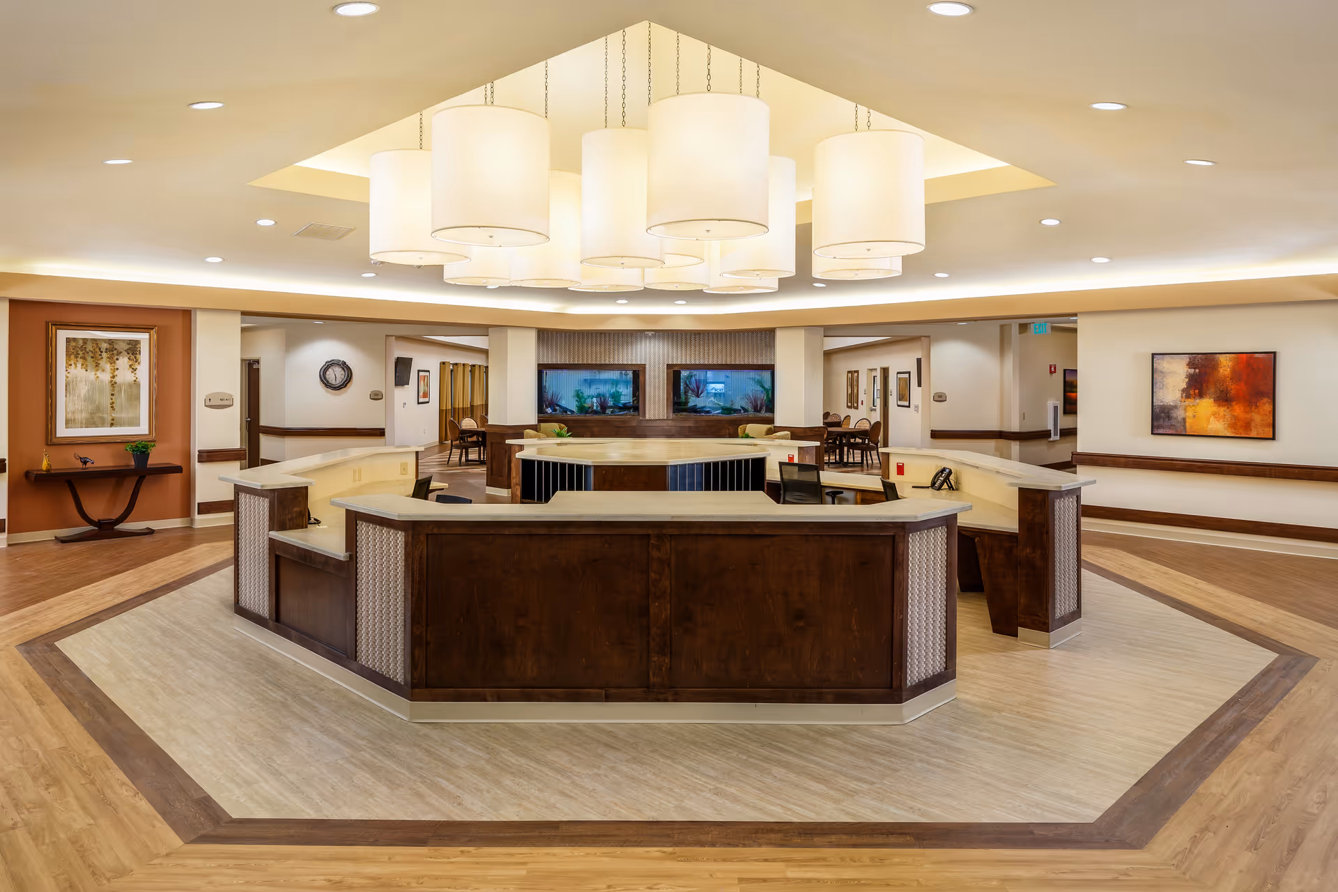 A spacious and well-lit reception area with a large, octagonal wooden front desk in the center. The ceiling features multiple large, cylindrical pendant lights. The floor has a wood and tile pattern, and the walls are decorated with framed artwork. There are hallways and seating areas visible in the background.