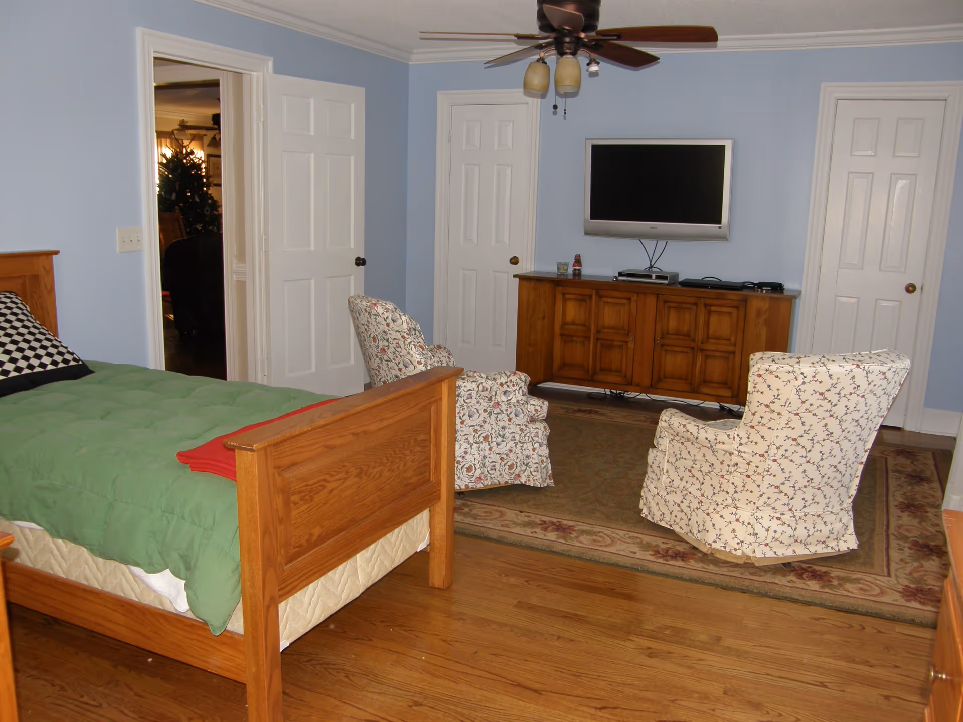 Spacious bedroom with a wooden bed, two patterned armchairs facing a wall-mounted TV above a wooden cabinet, a ceiling fan, and hardwood floors.