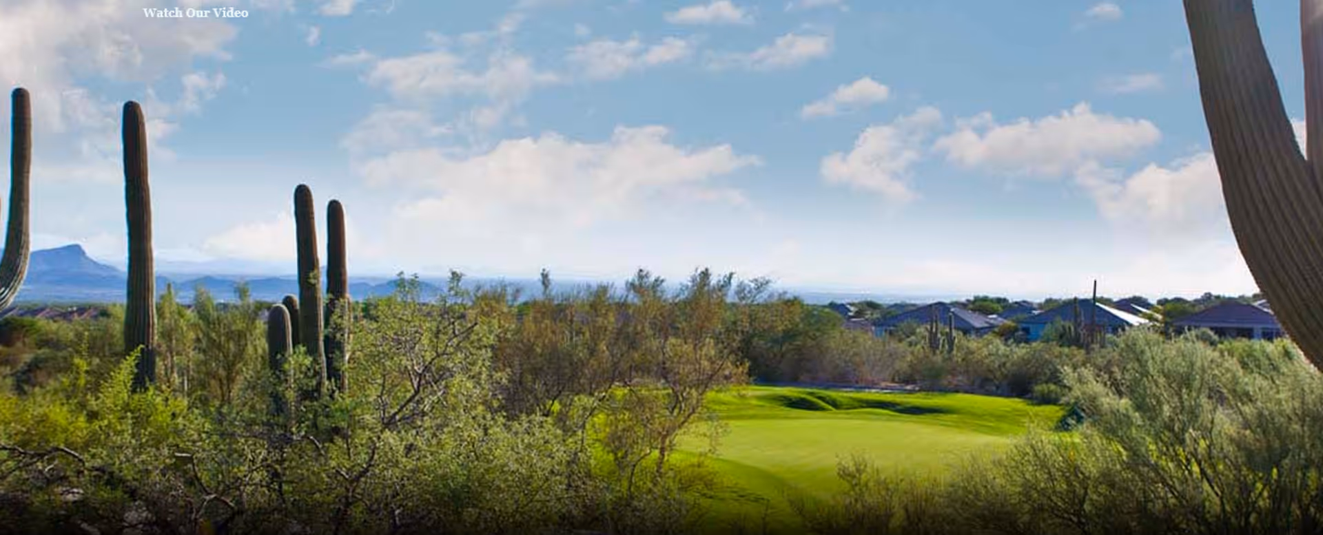Desert landscape with saguaro cacti overlooking a green golf course fairway and distant houses under a partly cloudy sky.