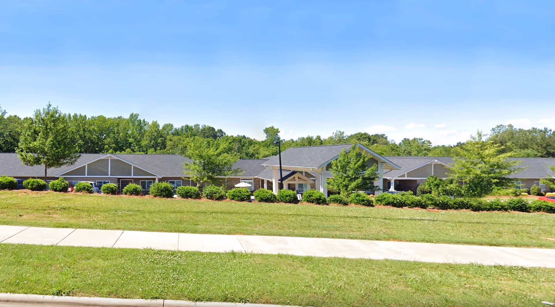 Exterior view of a single-story senior living facility building with a covered entrance, surrounded by green bushes, trees, and a well-maintained lawn under a clear blue sky.