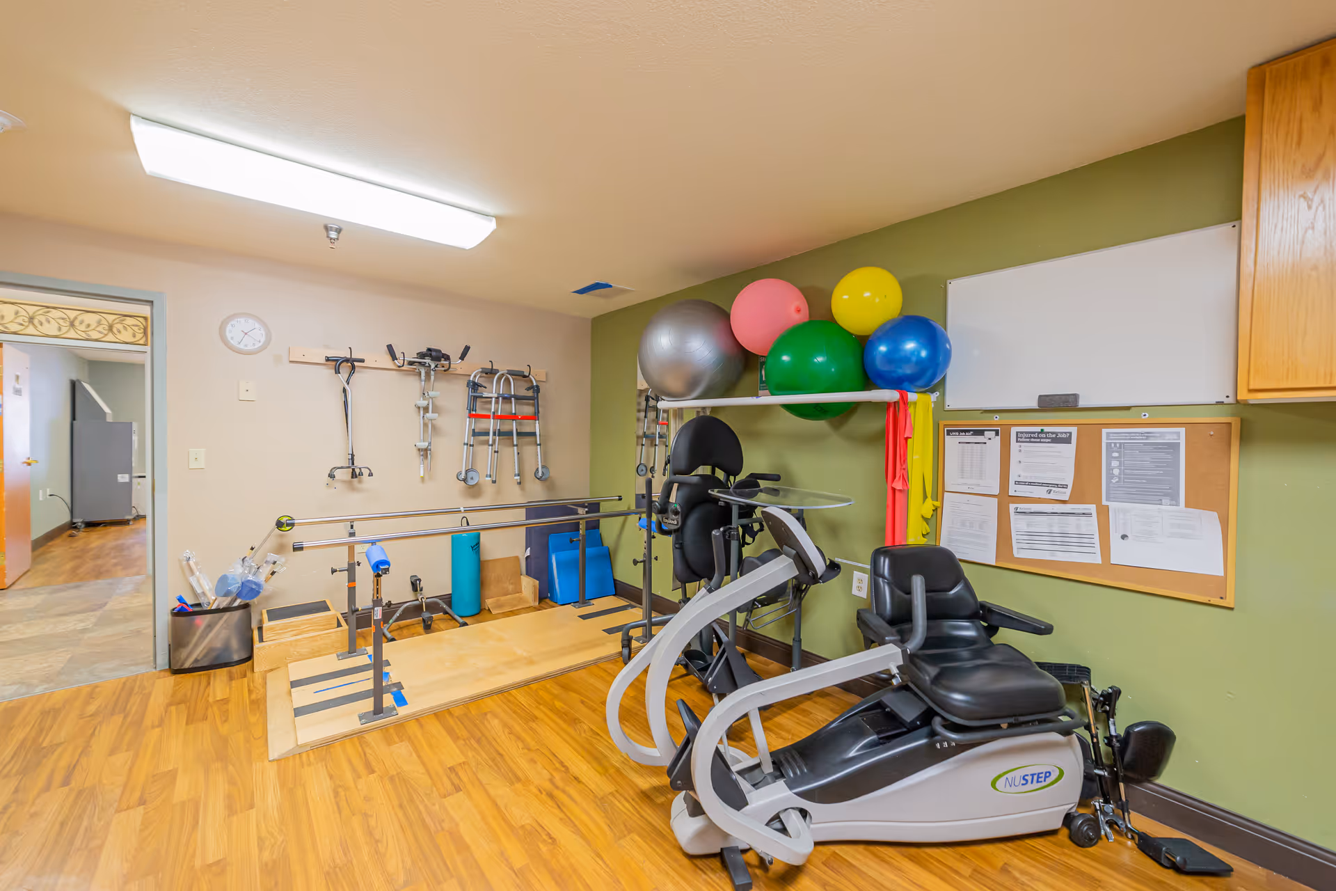 A physical therapy or rehabilitation room with exercise equipment including a NuStep recumbent cross trainer, parallel bars for walking practice, colorful exercise balls on a shelf, and various mobility aids such as walkers and crutches hanging on the wall. The room has wood flooring, a green accent wall with a whiteboard and bulletin board, and a doorway leading to another area.