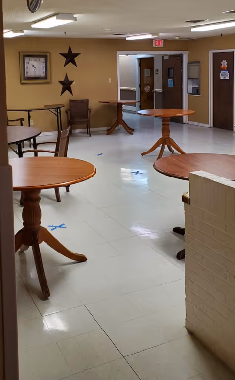 Interior view of a common area in a nursing home with several round wooden tables and chairs spaced apart on a tiled floor marked with blue tape Xs. The walls are beige with two decorative star wall hangings and a clock. There are double doors and other doors visible in the background.