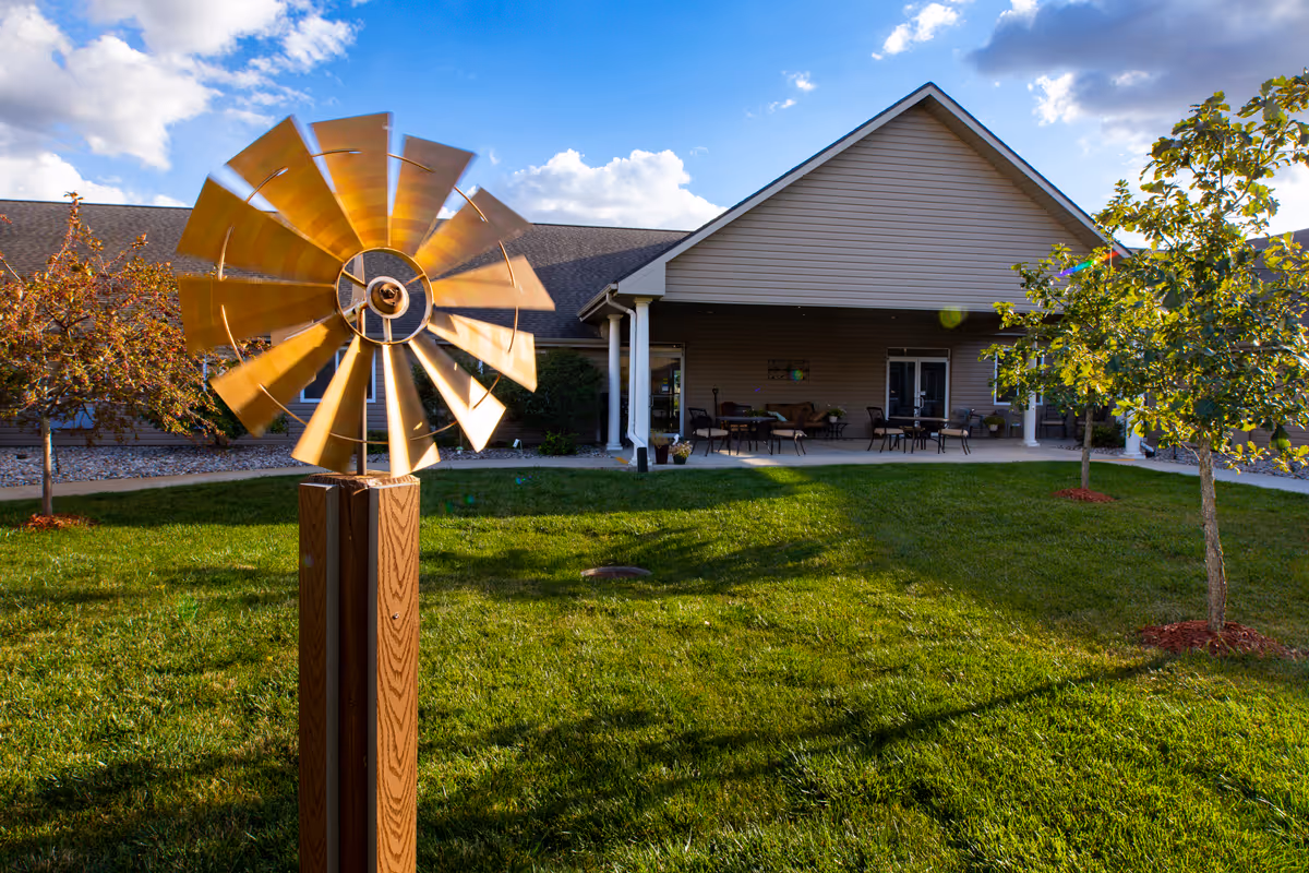 A decorative metal windmill on a grassy lawn in front of the entrance and covered patio of a senior living building.