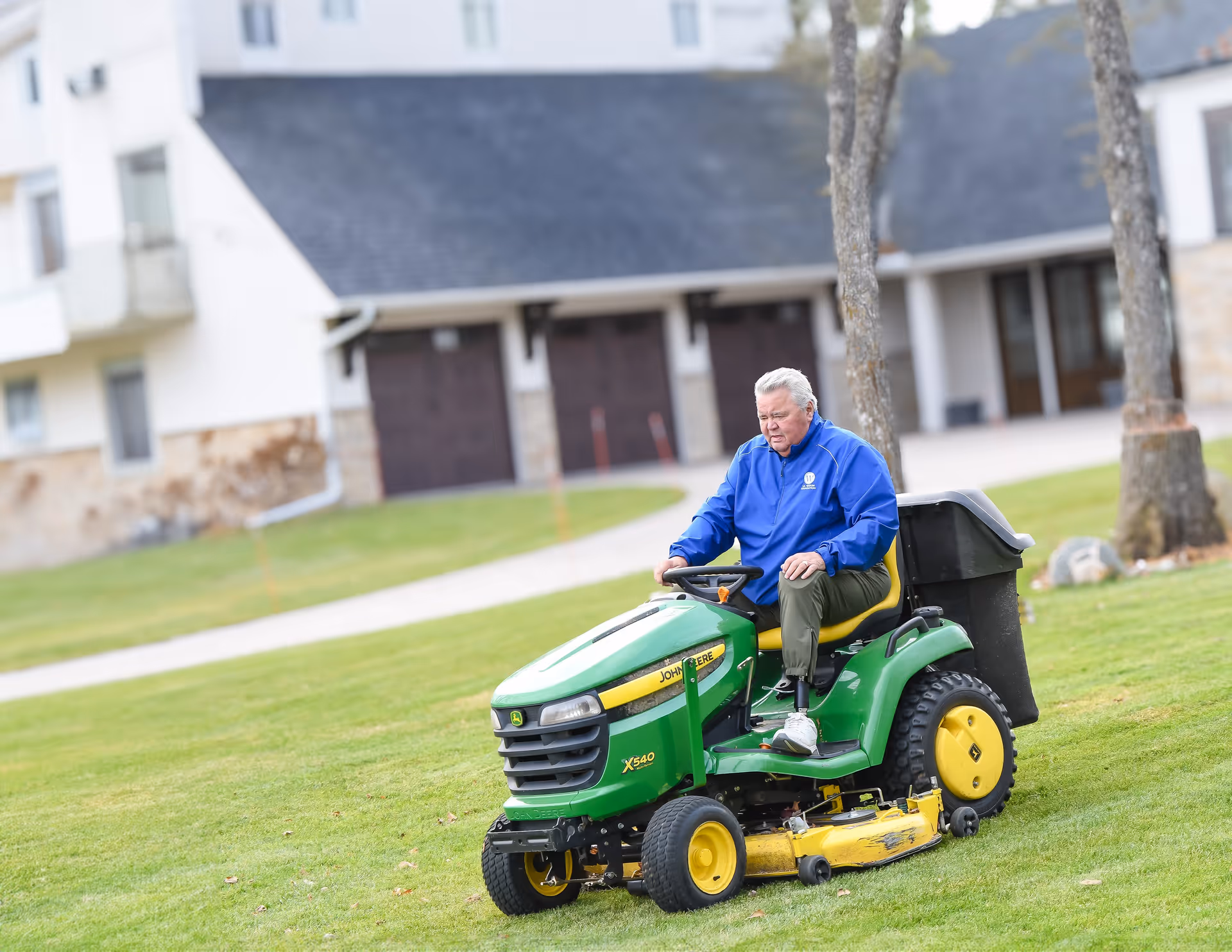 Older man drives a green John Deere mower across the grassy area outside Vivie Baxter.