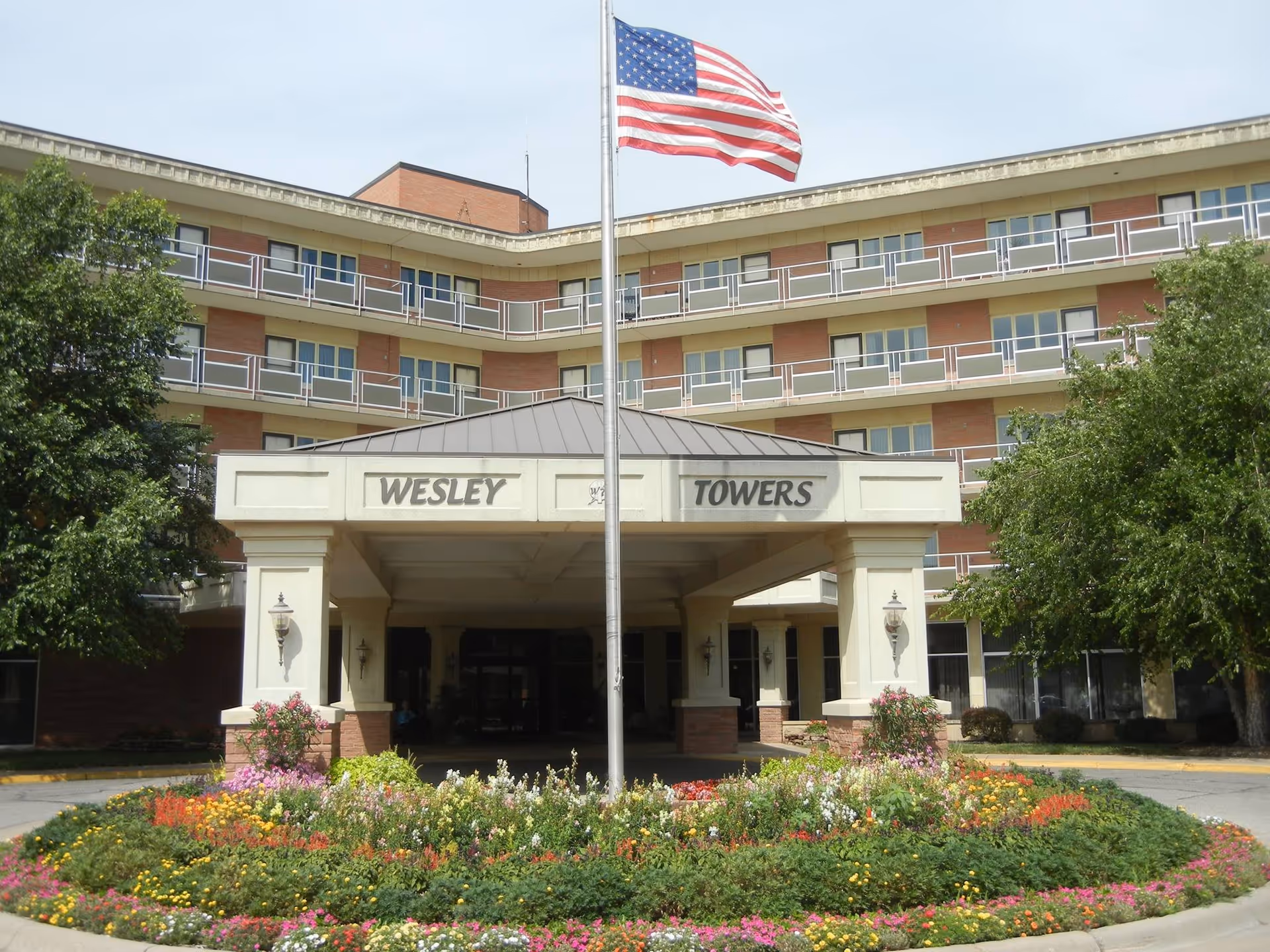 Front exterior view of Wesley Towers building with a covered entrance, an American flag on a flagpole, and a circular flower bed with colorful flowers in front.