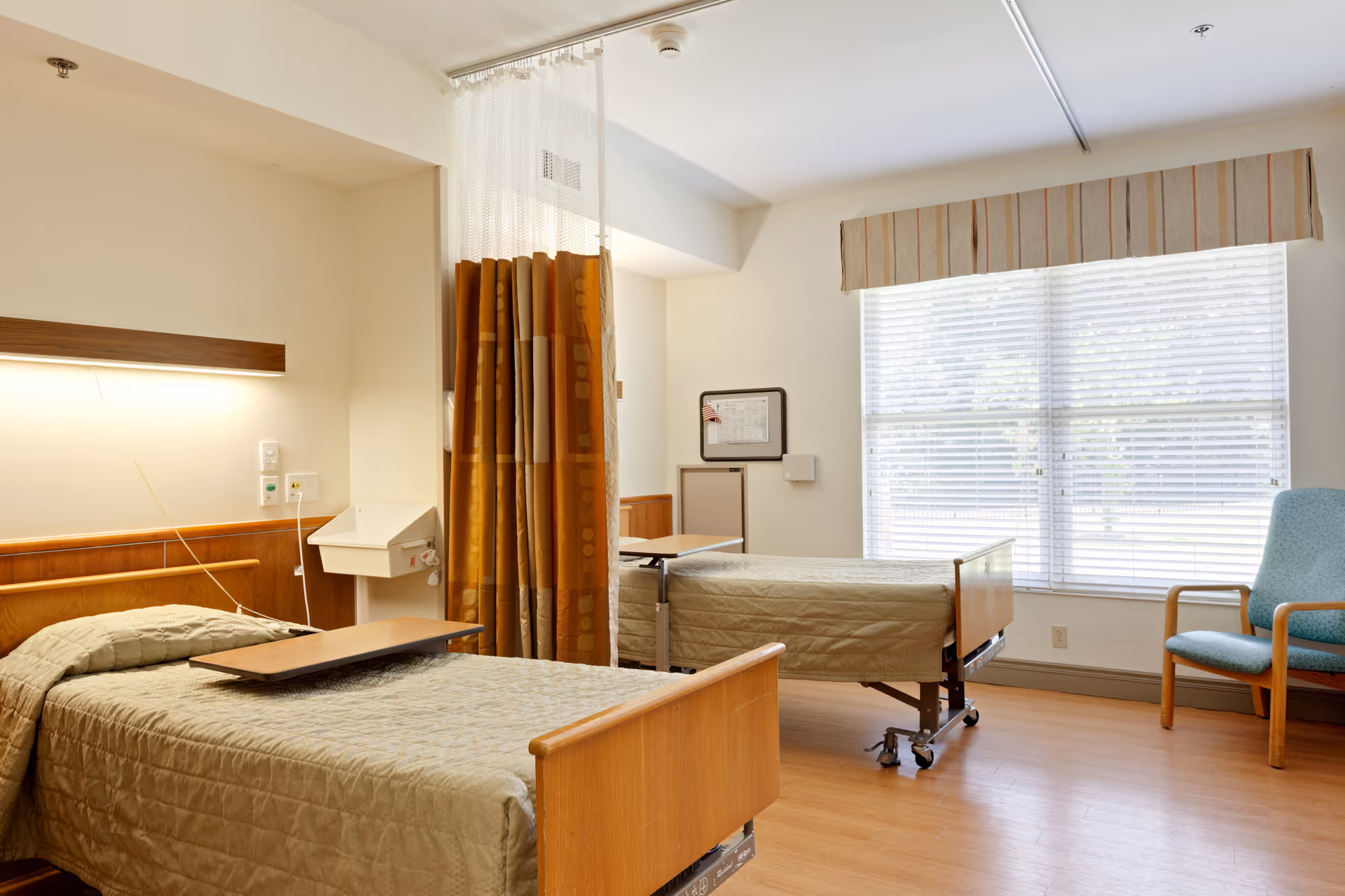 Two hospital beds separated by a privacy curtain in a bright patient room.