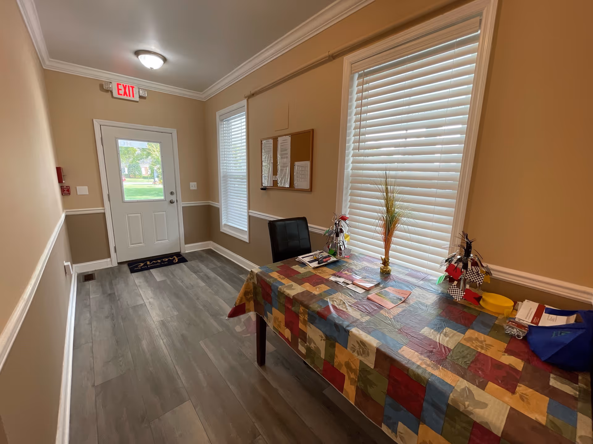 Small entry hallway with a door and two windows, a table covered with a colorful tablecloth holding brochures and decorations, and an EXIT sign above the door.