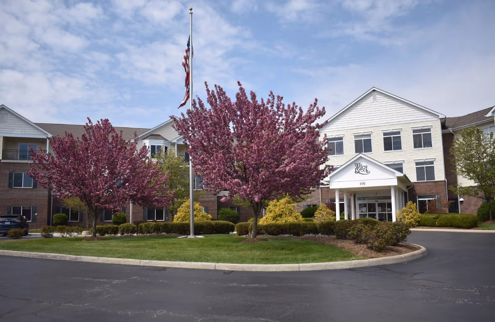 Front exterior view of Willow Brook at Delaware Run facility with a circular driveway, manicured lawn, blooming pink trees, an American flag on a flagpole, and a three-story building with white siding and brick accents under a partly cloudy sky.