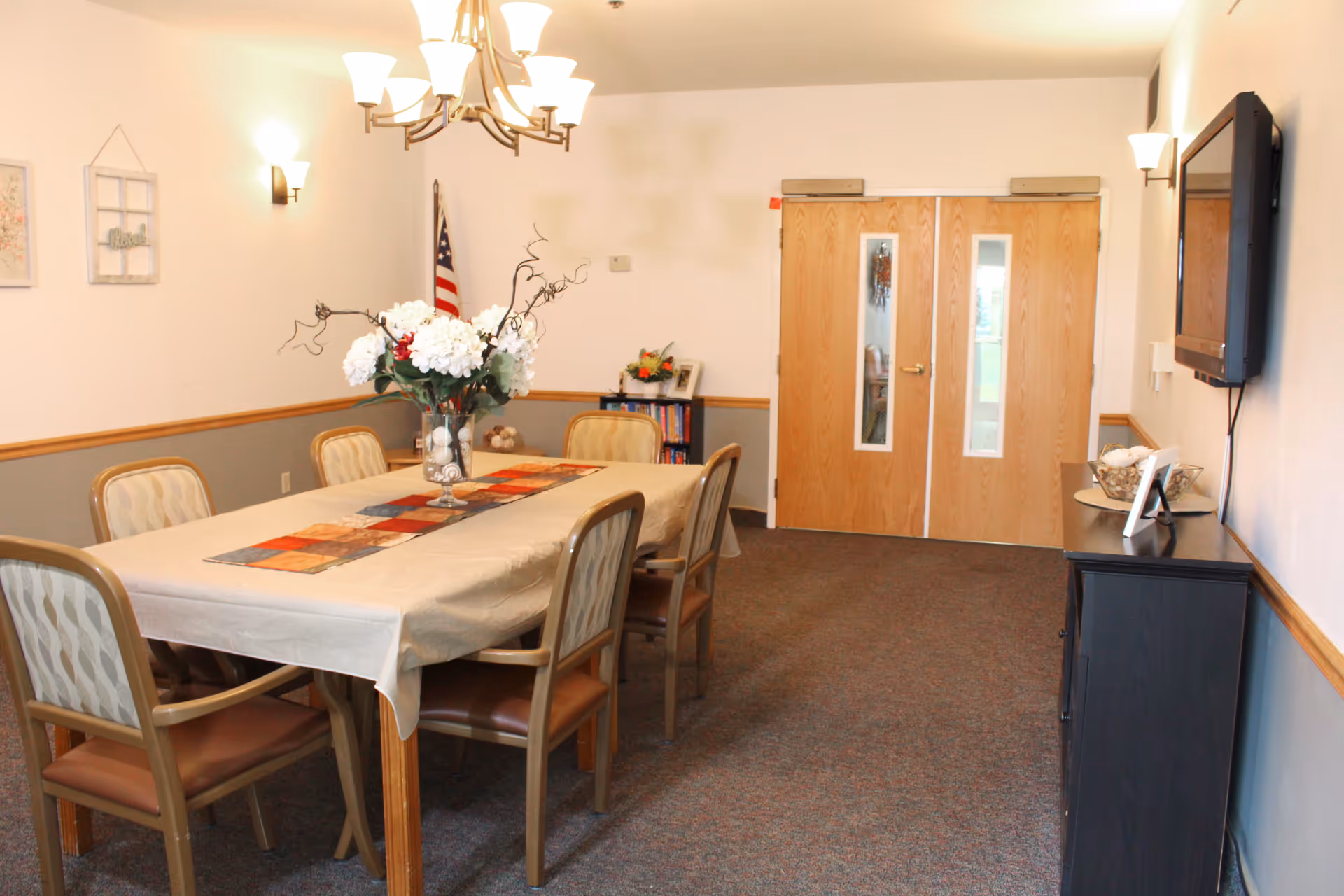 Bright dining room with a long table, chairs, chandelier, and cabinet in a senior living facility.