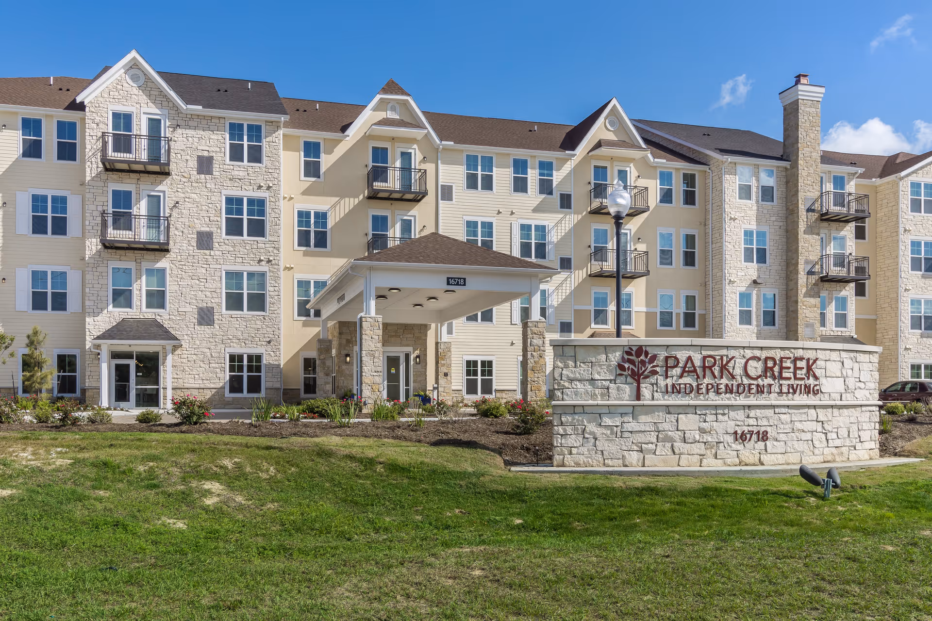 Exterior view of Park Creek Active Retirement building, a multi-story residential facility with beige and stone facade, balconies, a covered entrance, and a stone sign in front displaying the name and address 16718.