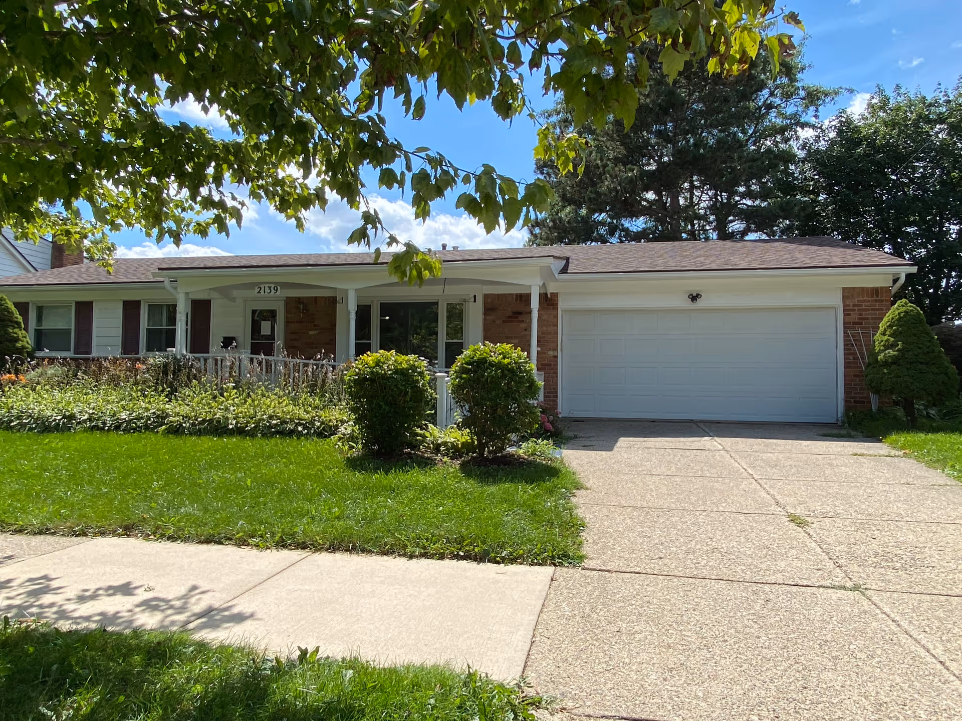 Single-story ranch-style house with an attached two-car garage, front porch, driveway and green lawn under leafy trees.