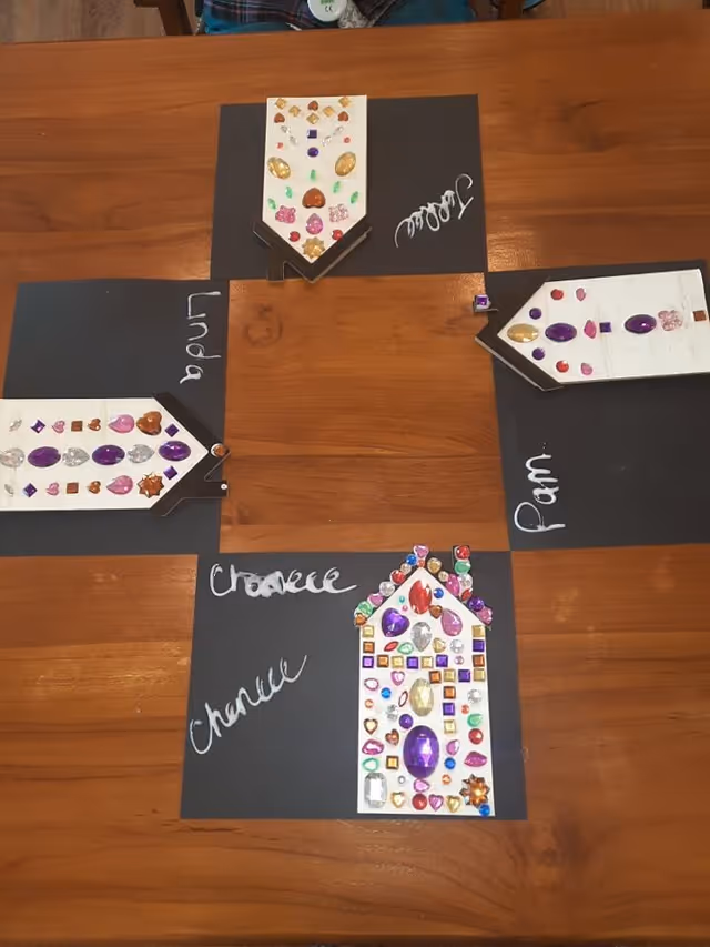 A wooden table with four black square papers arranged in a cross shape, each with a white paper house shape decorated with colorful gem stickers. Each black paper has a name written in white marker: Linda, Janice, Pam, and Charese.