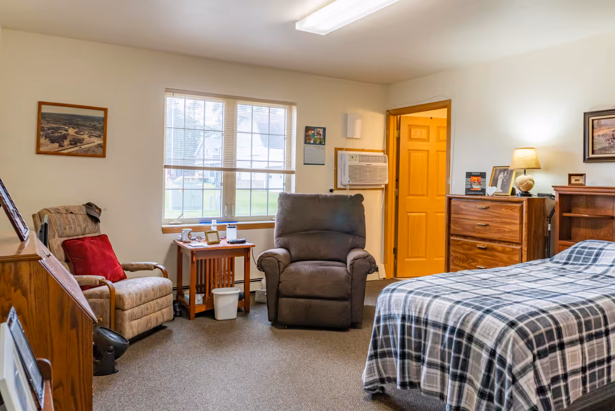 Private bedroom in a senior living facility with bed, recliner, dresser, and window.