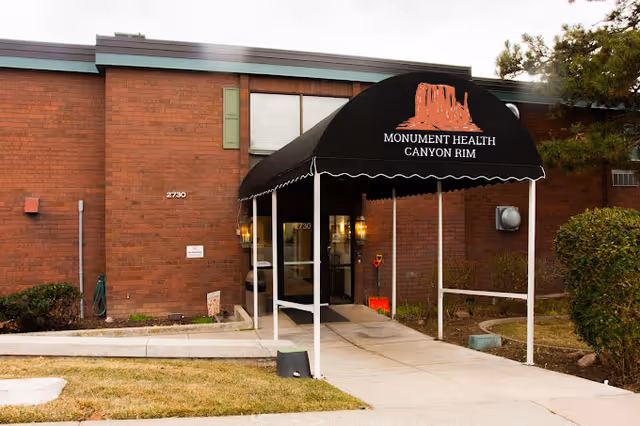 Entrance to Monument Health Canyon Rim facility with a black awning displaying the facility name and logo, a brick building, and a concrete walkway leading to glass doors.