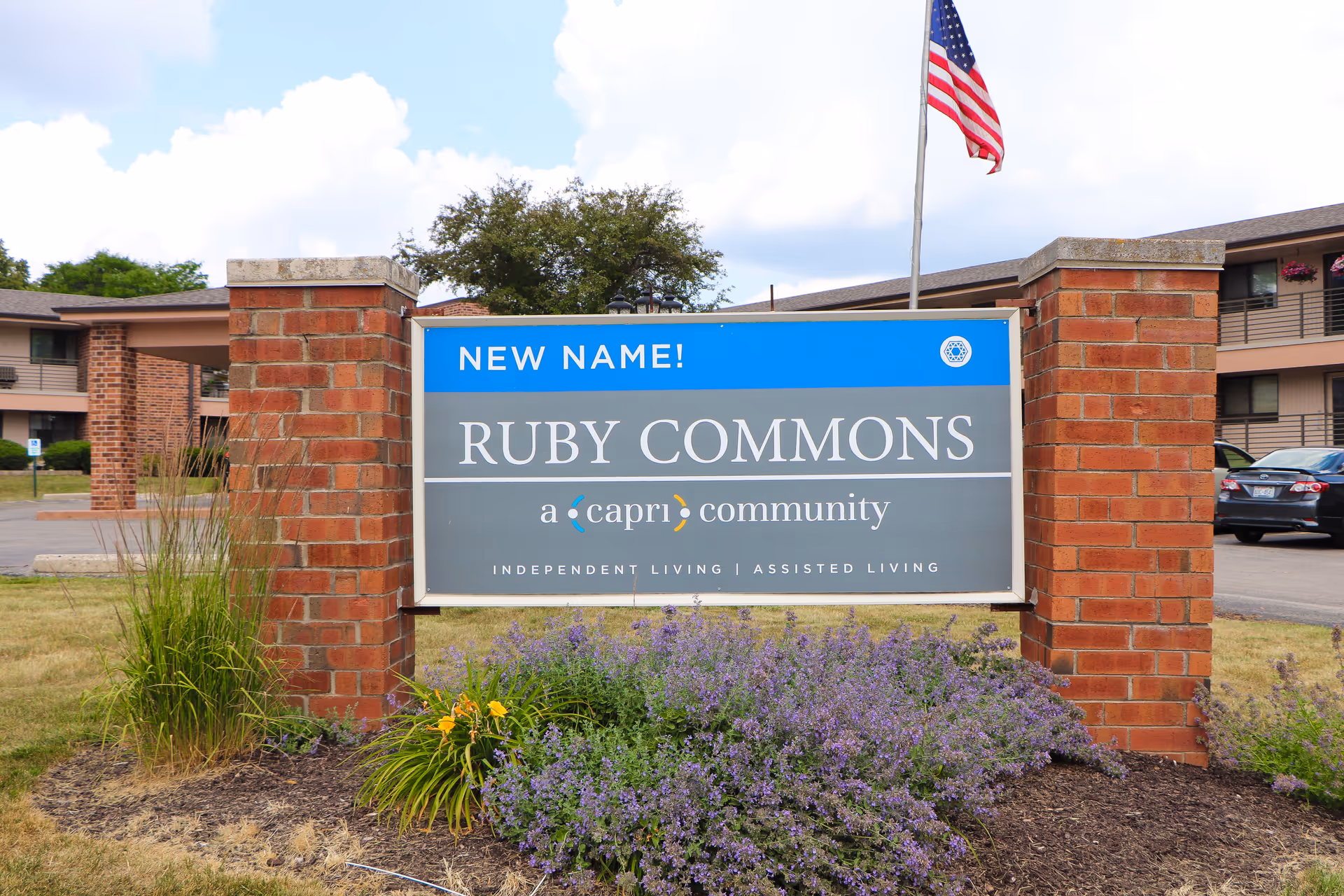 A large sign outside a senior living facility with brick pillars on each side. The sign reads 'NEW NAME! RUBY COMMONS a capri community INDEPENDENT LIVING | ASSISTED LIVING'. There are purple flowers and green plants in front of the sign, and an American flag is visible in the background along with parts of the building and parked cars.