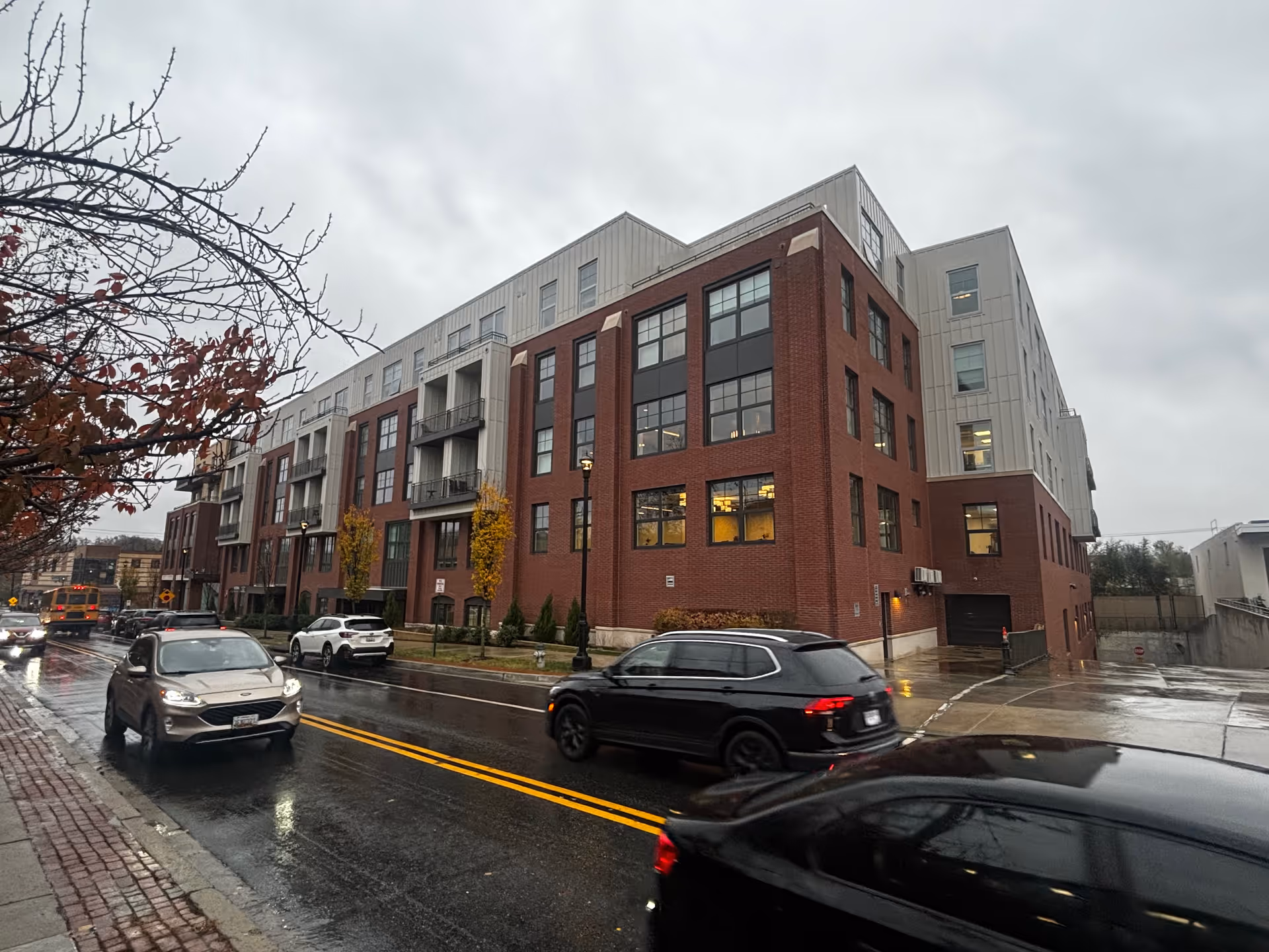 Five-story brick-and metal-clad apartment/assisted-living building along a wet city street with cars and bare trees under an overcast sky.