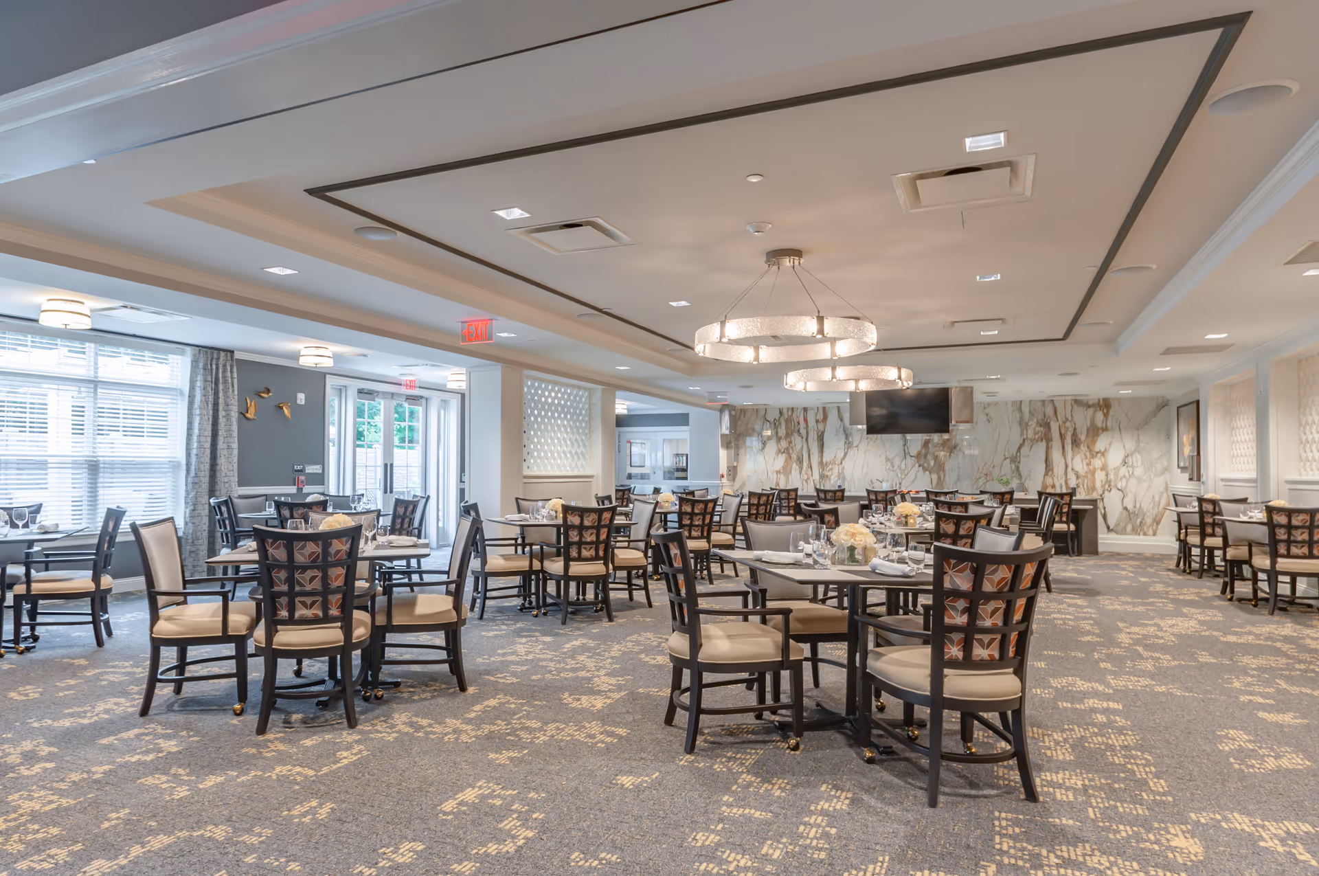 A spacious and well-lit dining room with multiple round tables and cushioned chairs arranged neatly. The room features large windows with curtains allowing natural light, elegant chandeliers hanging from the ceiling, and a marble accent wall with a mounted flat-screen TV. The carpet has a patterned design, and the overall decor is modern and inviting.