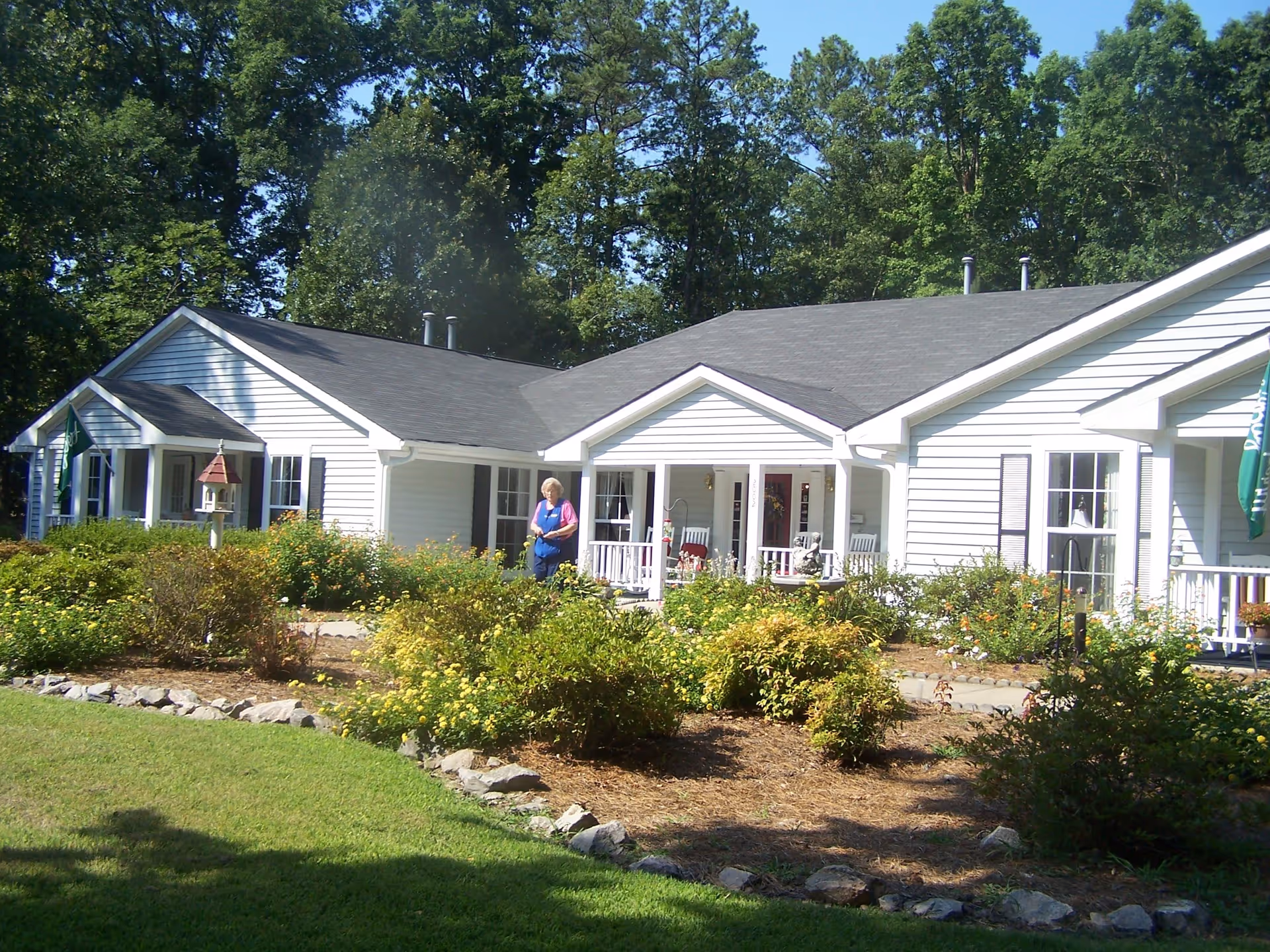 Exterior view of a white single-story building with a dark roof, surrounded by green bushes and trees. A person is standing near the porch area, which has white railings and rocking chairs. The sky is clear and blue.