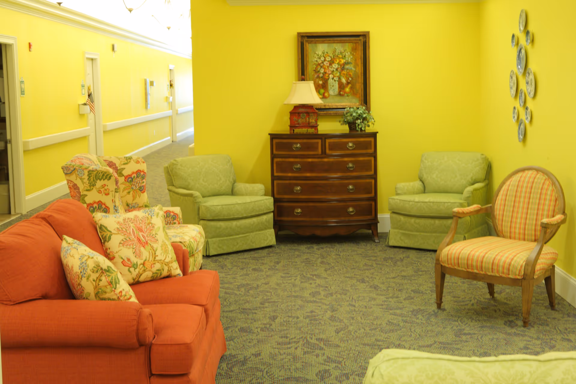 A cozy sitting area in a senior living facility with bright yellow walls, featuring a red sofa with floral cushions, two green armchairs, a striped armchair, a wooden chest of drawers with a lamp and a plant on top, and a framed floral painting on the wall. The carpet is patterned, and there is a hallway with doors visible in the background.