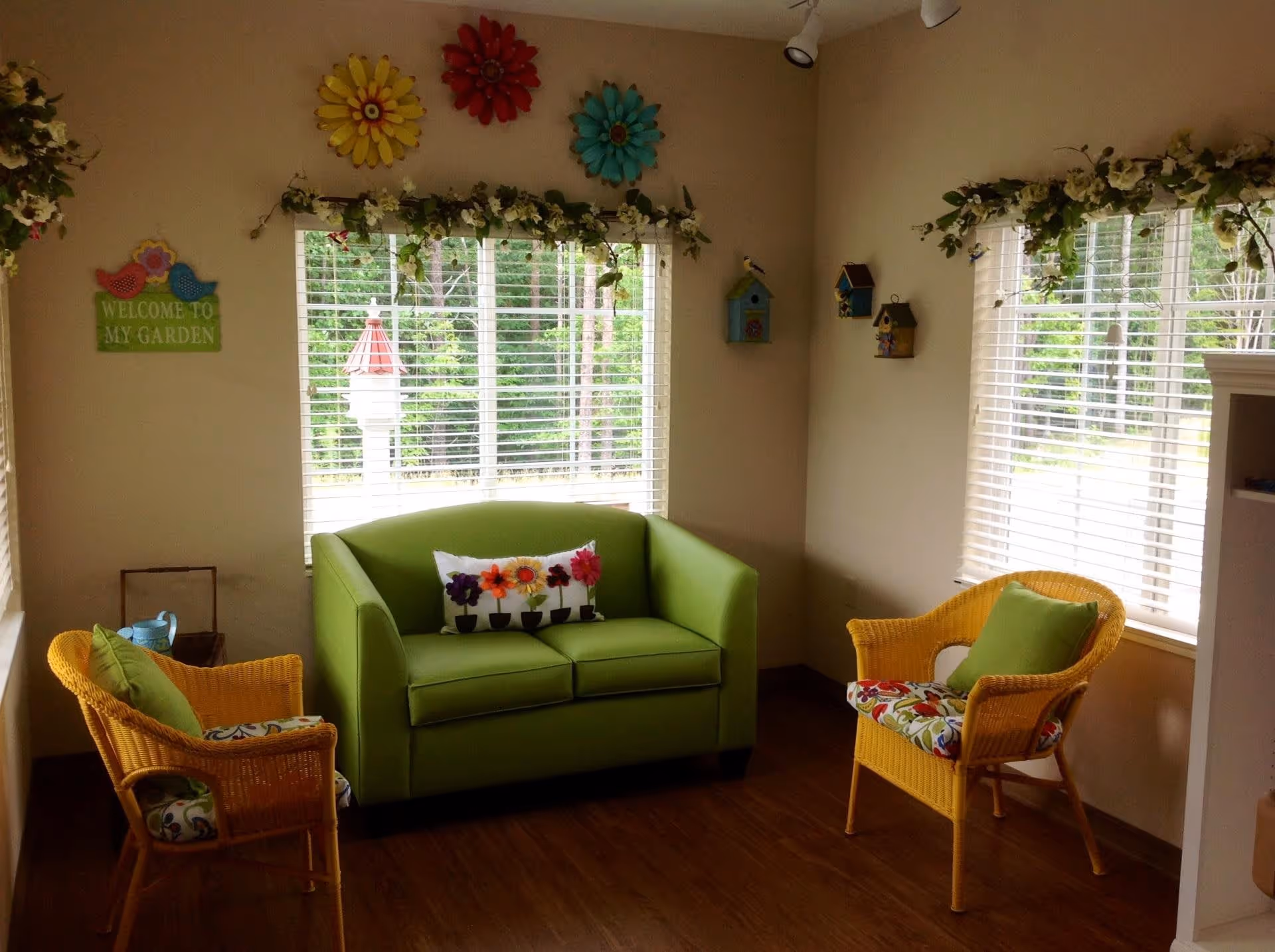 A cozy sitting area with a green loveseat and two yellow wicker chairs with floral cushions. The room has large windows with white blinds, decorated with garlands of flowers. Colorful flower wall decorations and small birdhouses adorn the walls. A sign on the wall reads 'Welcome to my garden.'