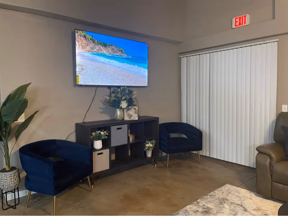 A small common living area with a wall-mounted TV, two navy armchairs, a console with decor, and vertical blinds.