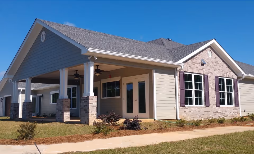 Exterior view of a single-story building with a covered porch supported by white columns with brick bases. The building has beige siding, a gray shingled roof, and multiple windows with purple shutters. There is a concrete walkway and some landscaping with small bushes in front of the building under a clear blue sky.