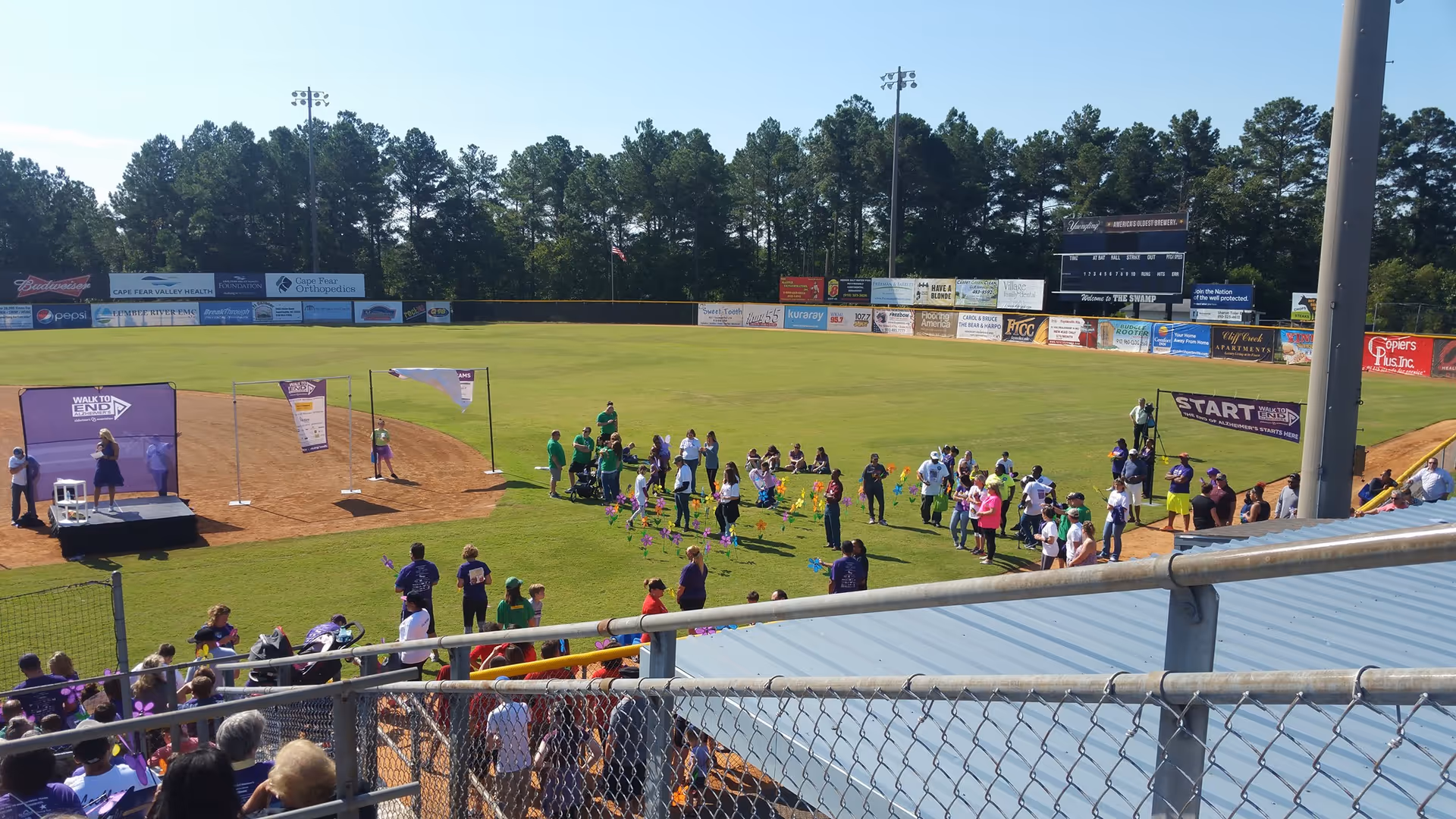 Sunny outdoor baseball field with a crowd gathered around a stage and banners.