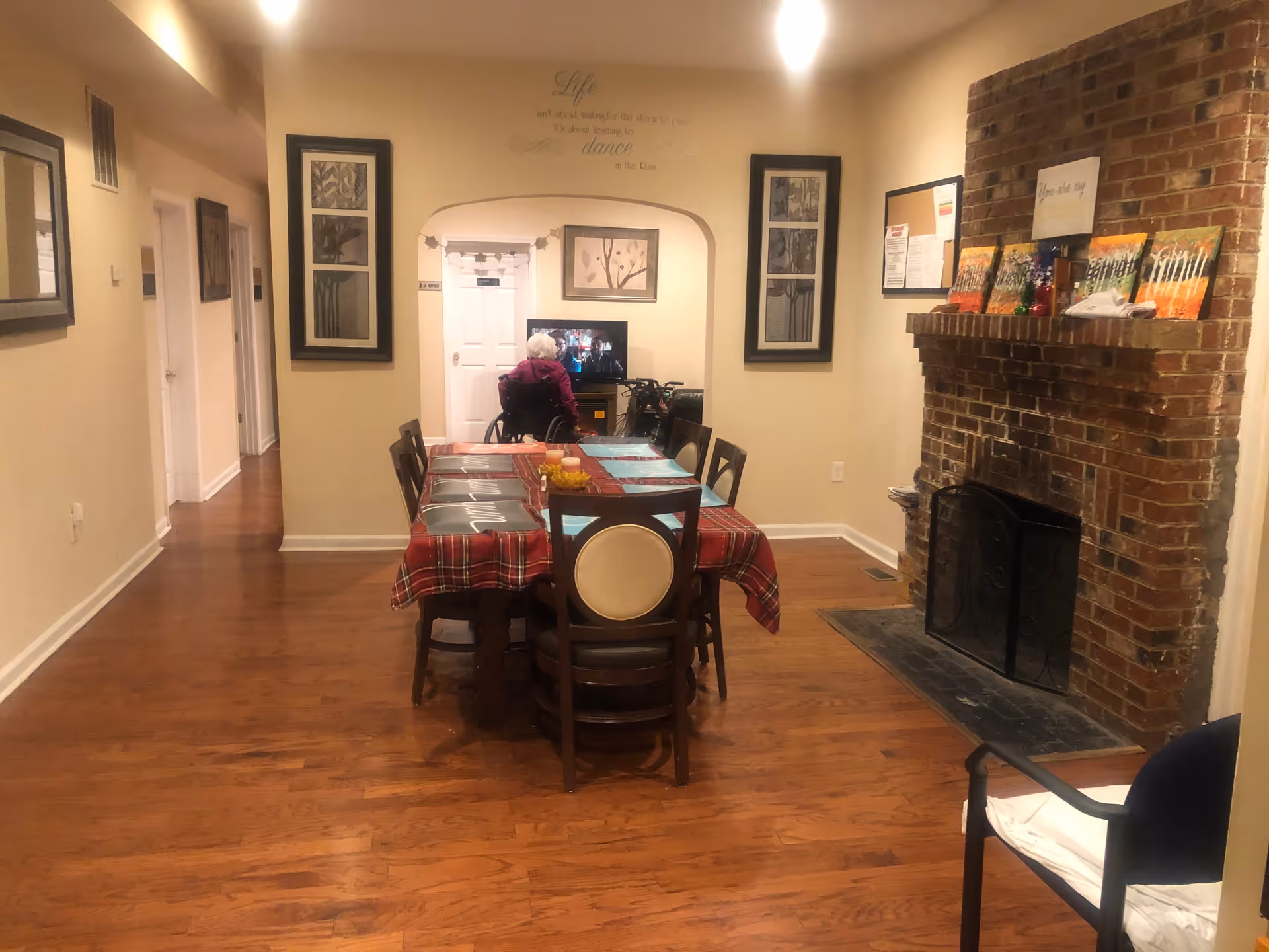 A dining room with a long table covered by a red plaid tablecloth and placemats, surrounded by wooden chairs. A brick fireplace is on the right wall with decorative items on the mantel. In the background, an elderly person in a wheelchair is watching a television in an adjacent room. The walls are decorated with framed artwork and a motivational quote above the doorway.