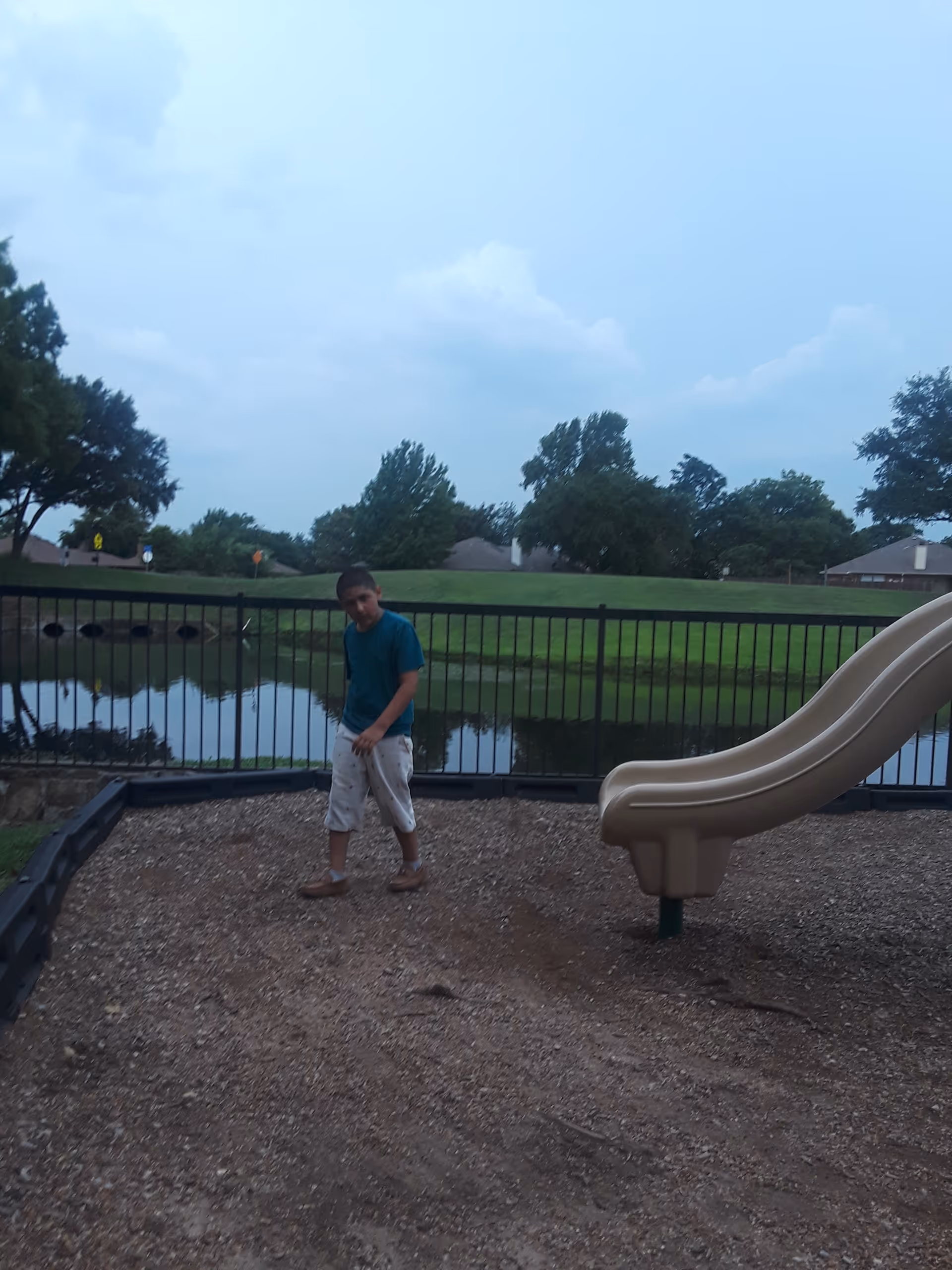 A person walks on a gravel playground near a beige slide by a pond.