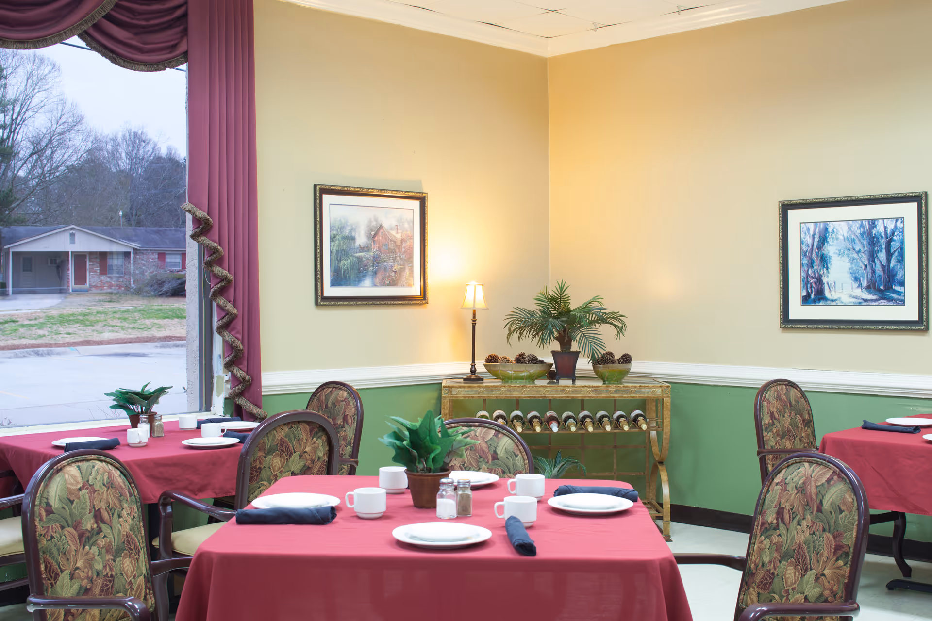 Dining room with several tables covered in red tablecloths set with plates and cups, upholstered chairs, framed paintings on the walls and a sideboard with plants and wine storage.