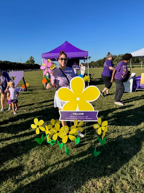A woman stands outdoors on a grassy field with purple tents and oversized yellow flower signs.