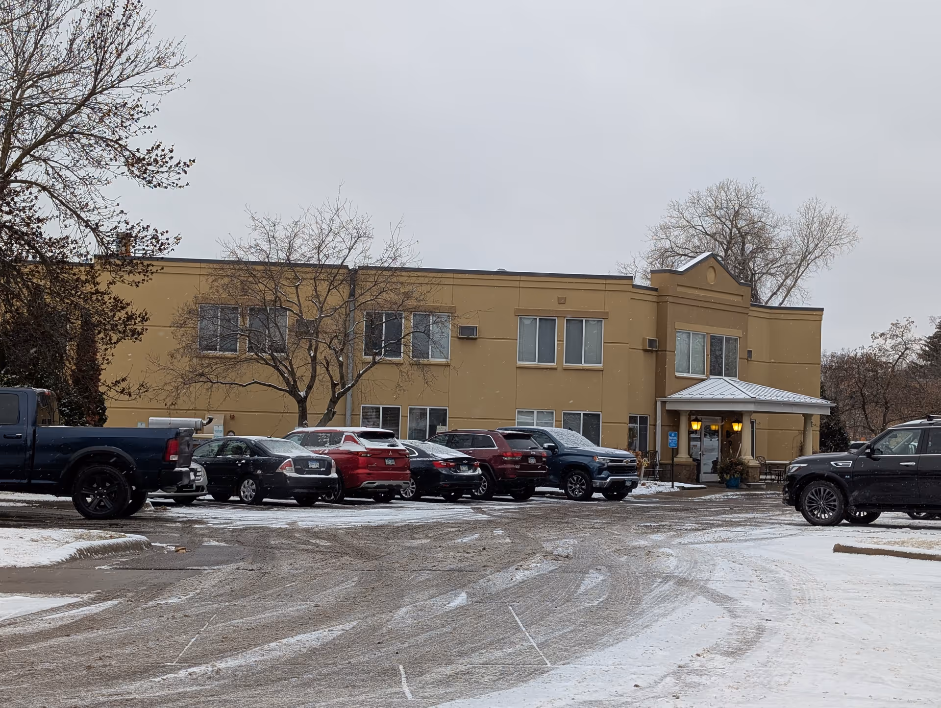 Two-story beige building with a covered entrance, parked cars in front, and light snow on the driveway.