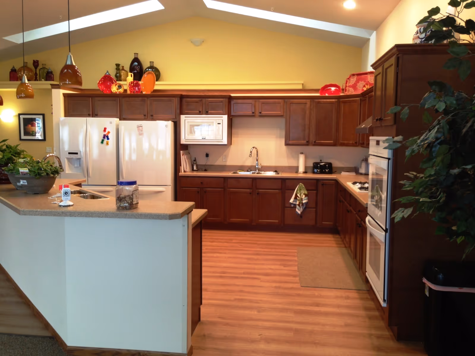 A spacious kitchen with wooden cabinets, a white refrigerator, a microwave, a double oven, and a sink. The kitchen has a beige countertop with a small plant and a jar on it. The walls are painted yellow, and there are decorative glass bottles on top of the cabinets. The floor is wooden, and there is a small mat in front of the oven. Pendant lights hang from the ceiling.